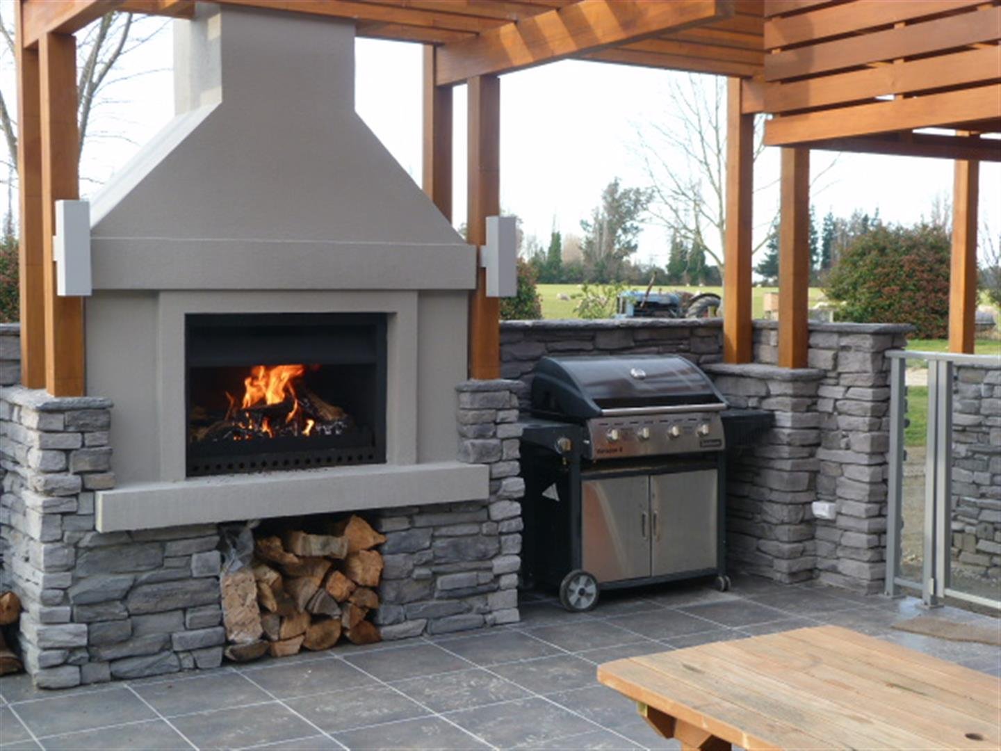 Outdoor patio area with a fireplace, wood rack, and gas grill, surrounded by a stone wall and wooden pergola, overlooking a green field with trees in the background.