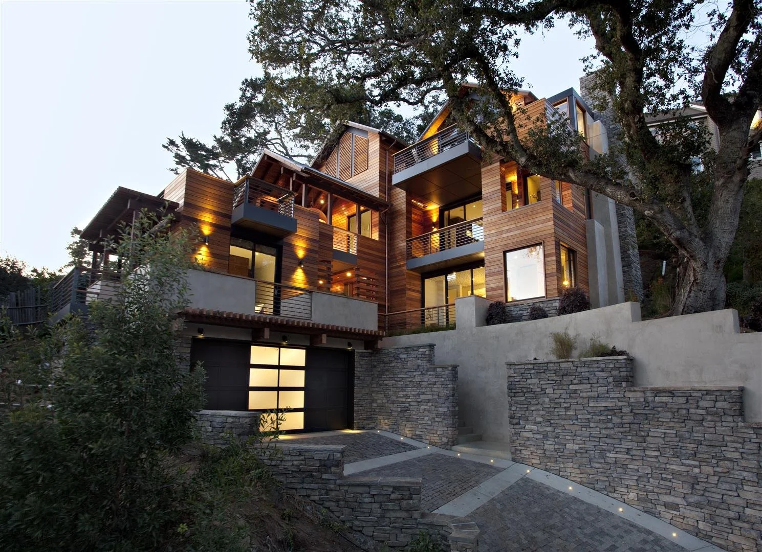 Modern multi-story house with large windows and wooden siding, built on a hillside with stone retaining walls and a paved driveway, surrounded by trees at dusk.