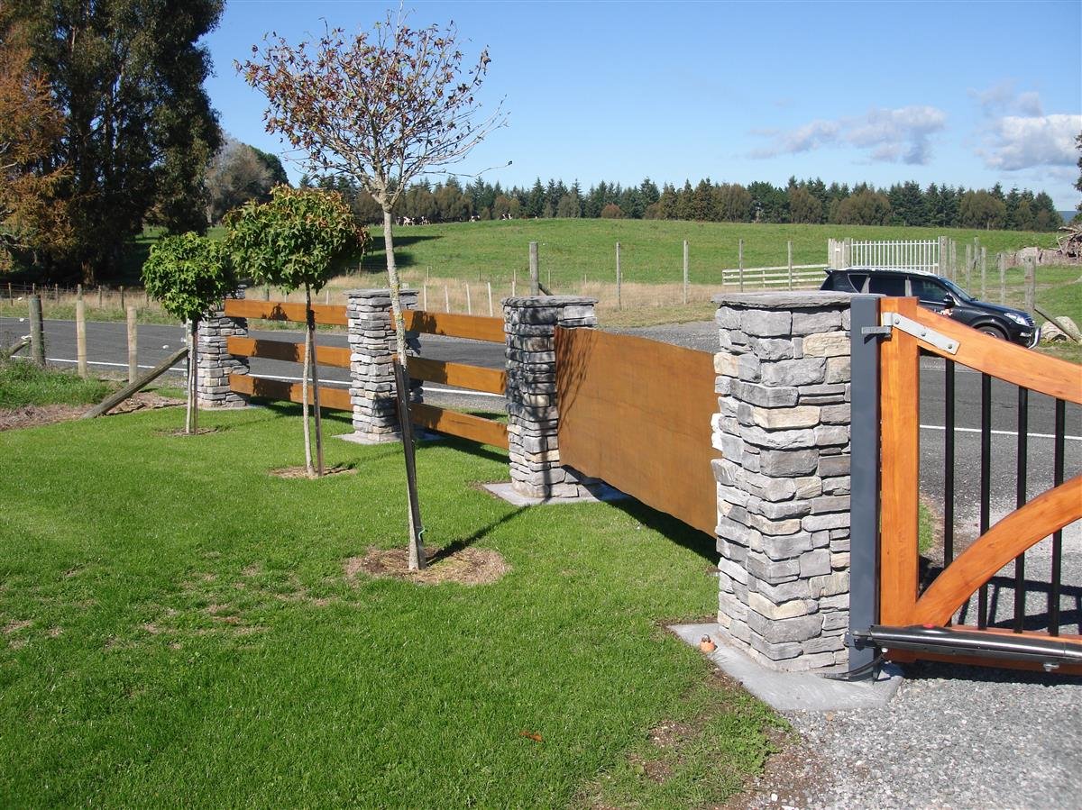 A decorative fence with stone pillars, wooden and metal panels, surrounding a grassy area with small trees, adjacent to a paved parking lot with a black SUV, under a clear blue sky.