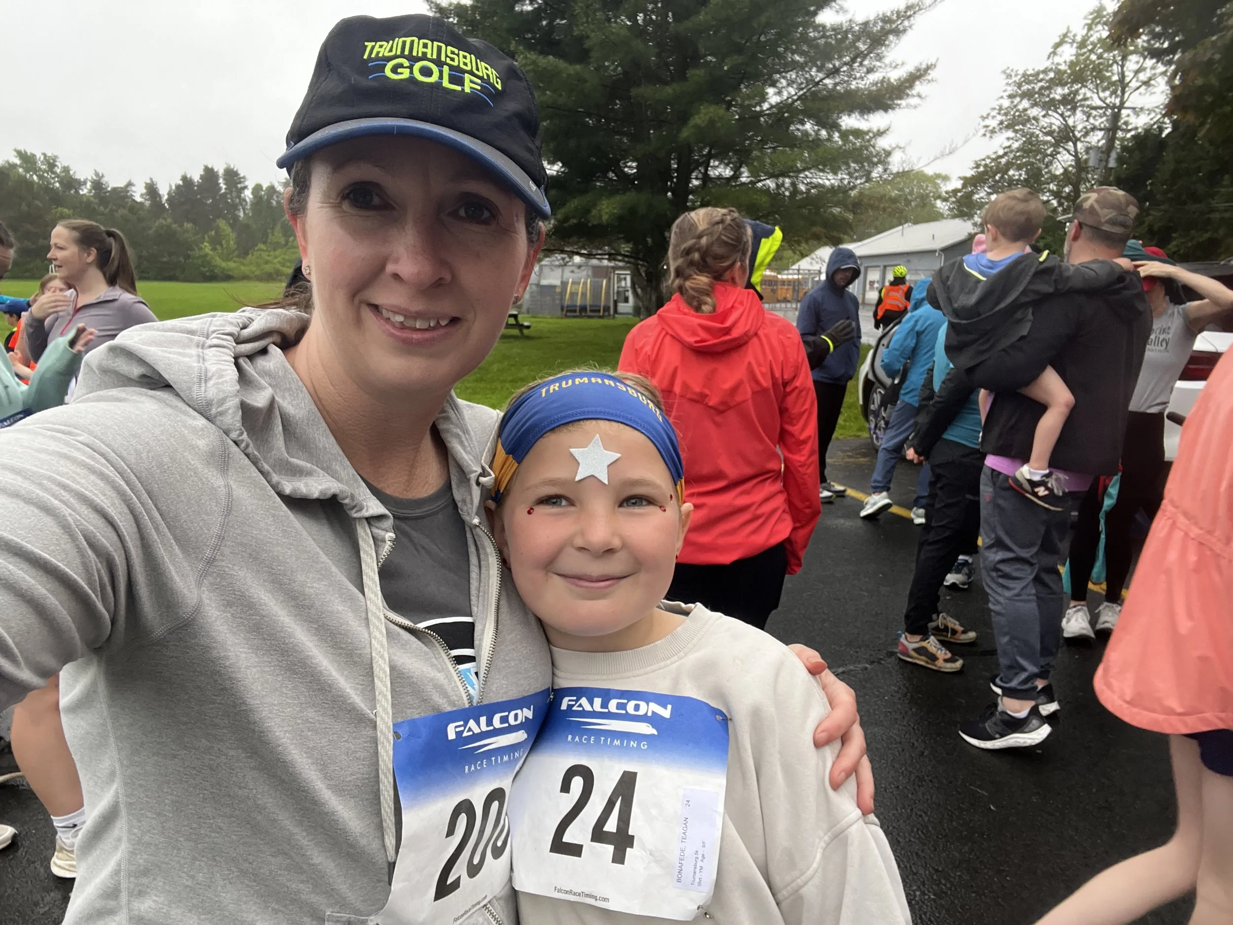Katelin and her daughter at a 5K race, both wearing race bibs and smiling. Katelin is wearing a black cap with yellow lettering and a gray jacket; her daughterl has a star sticker on her forehead, a blue headband, and a white sweatshirt.