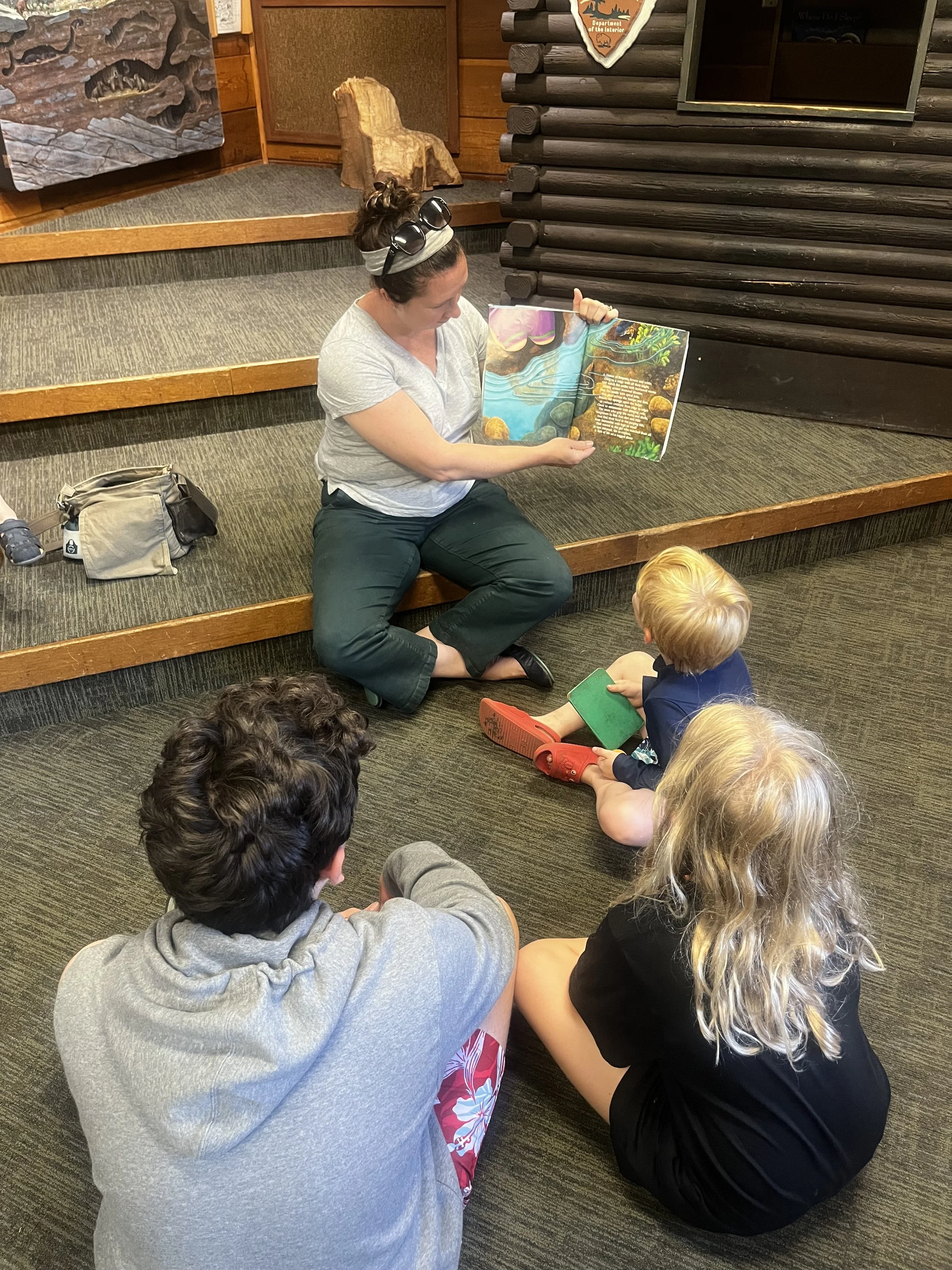 Katelin reading a colorful picture book to three children sitting on the floor.