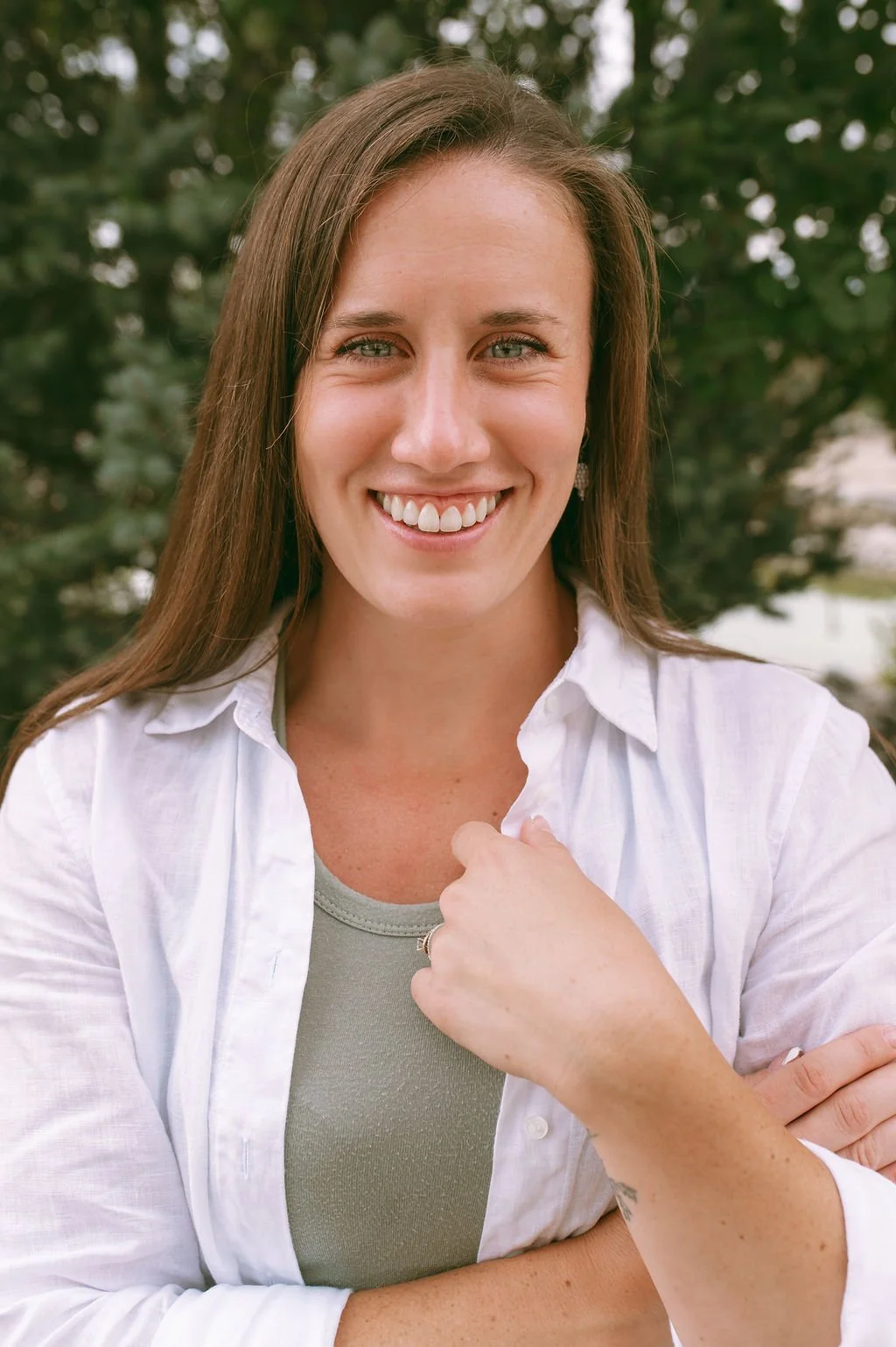 A woman with long brown hair and blue eyes smiling outdoors, wearing a white button-up shirt over a gray top, with a background of green trees.