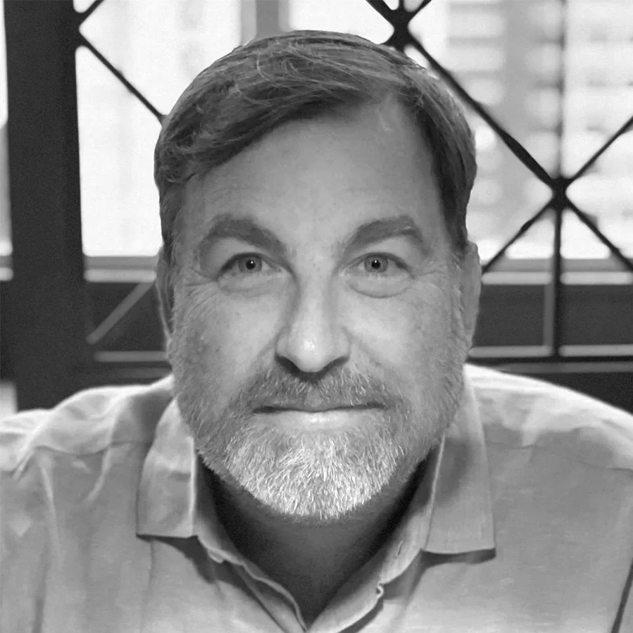 Black and white photo of a middle-aged man with a beard, smiling at the camera, in an indoor setting with a window and patterned grid in the background.