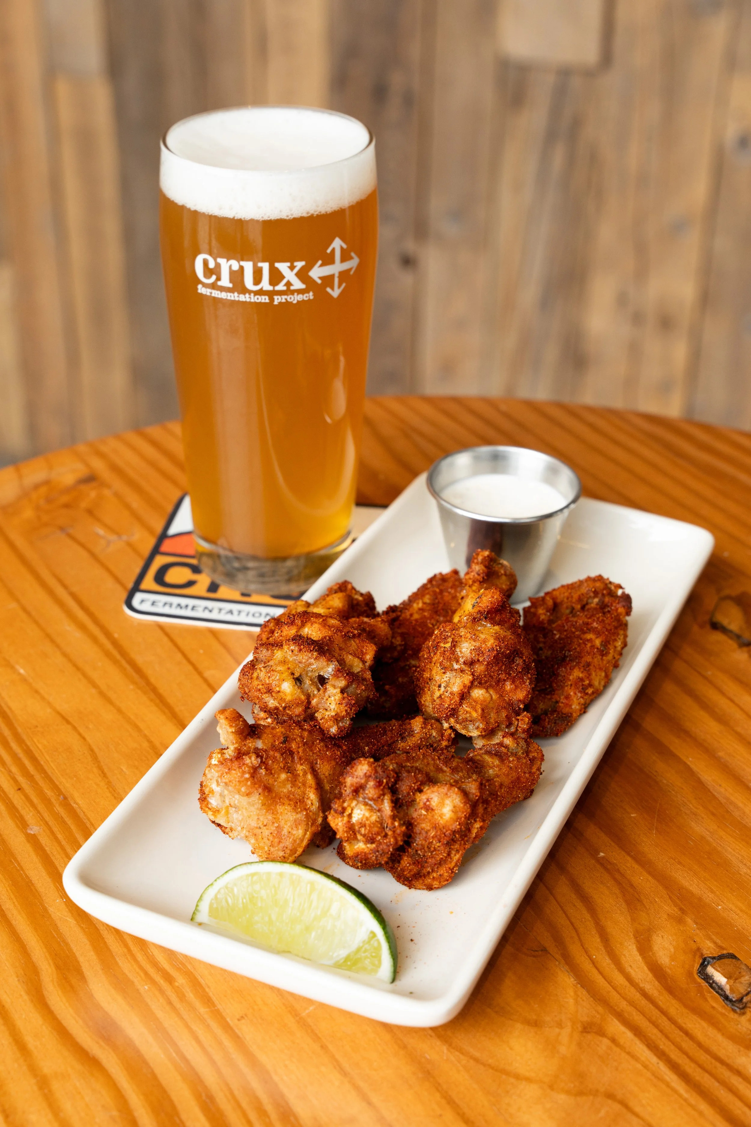 A pint glass of beer, a plate of fried chicken wings with a lemon wedge, and a small container of dipping sauce on a wooden table.