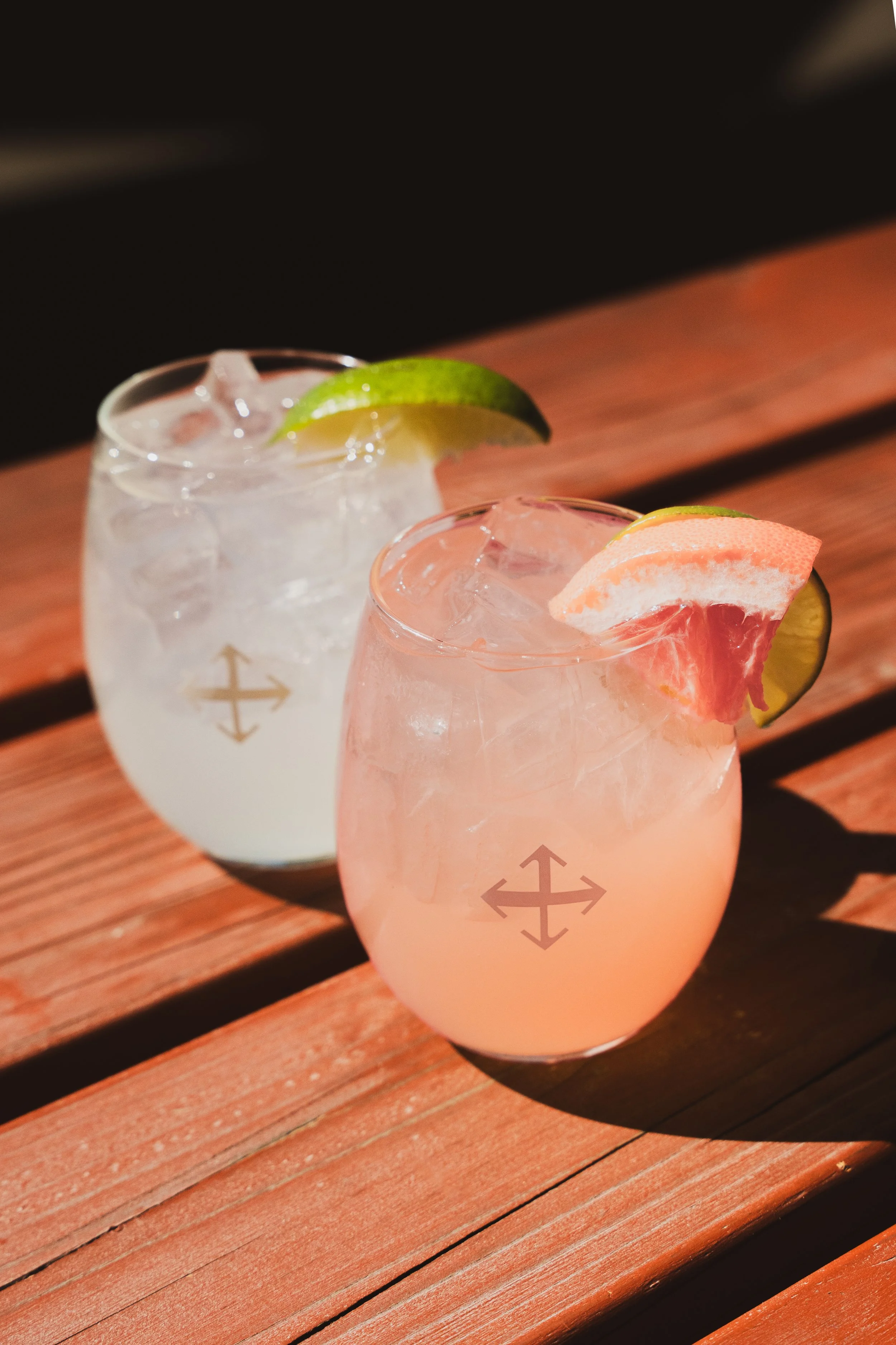 Two colorful cocktails with ice, lime, and grapefruit garnishes on a wooden table.