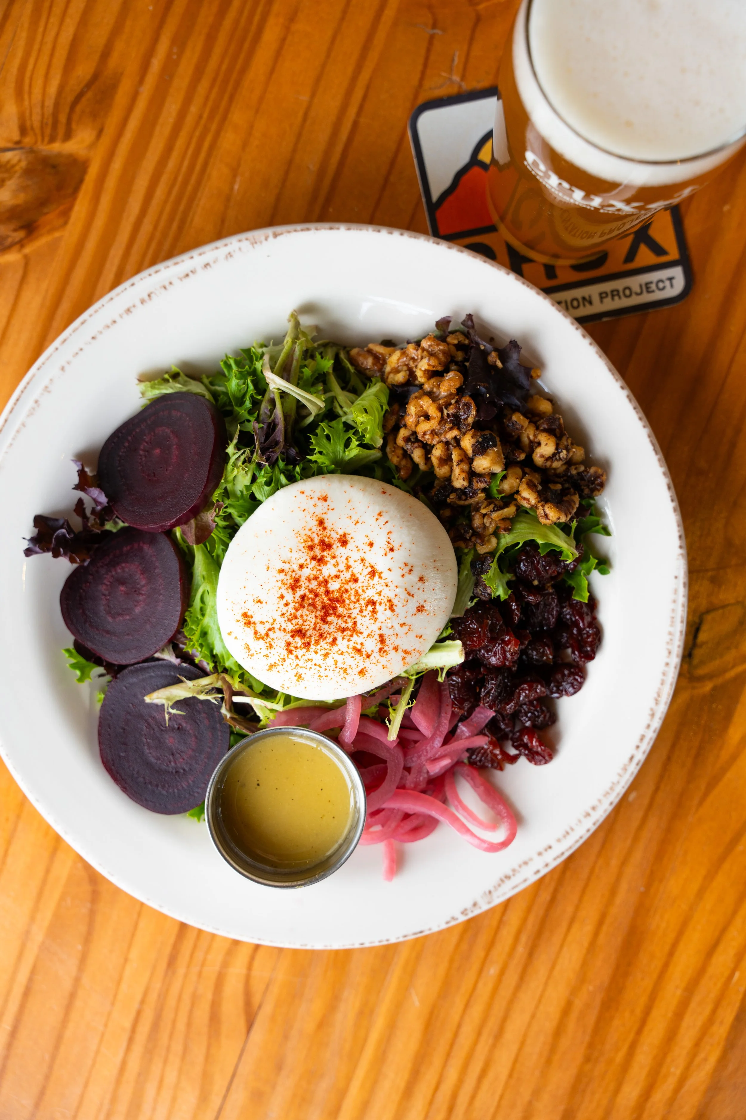 A salad with beet slices, mixed greens, shredded pickled onions, dried cranberries, a soft-boiled egg sprinkled with paprika, chopped nuts, and a small container of dressing, with a glass of beer on a wooden table.