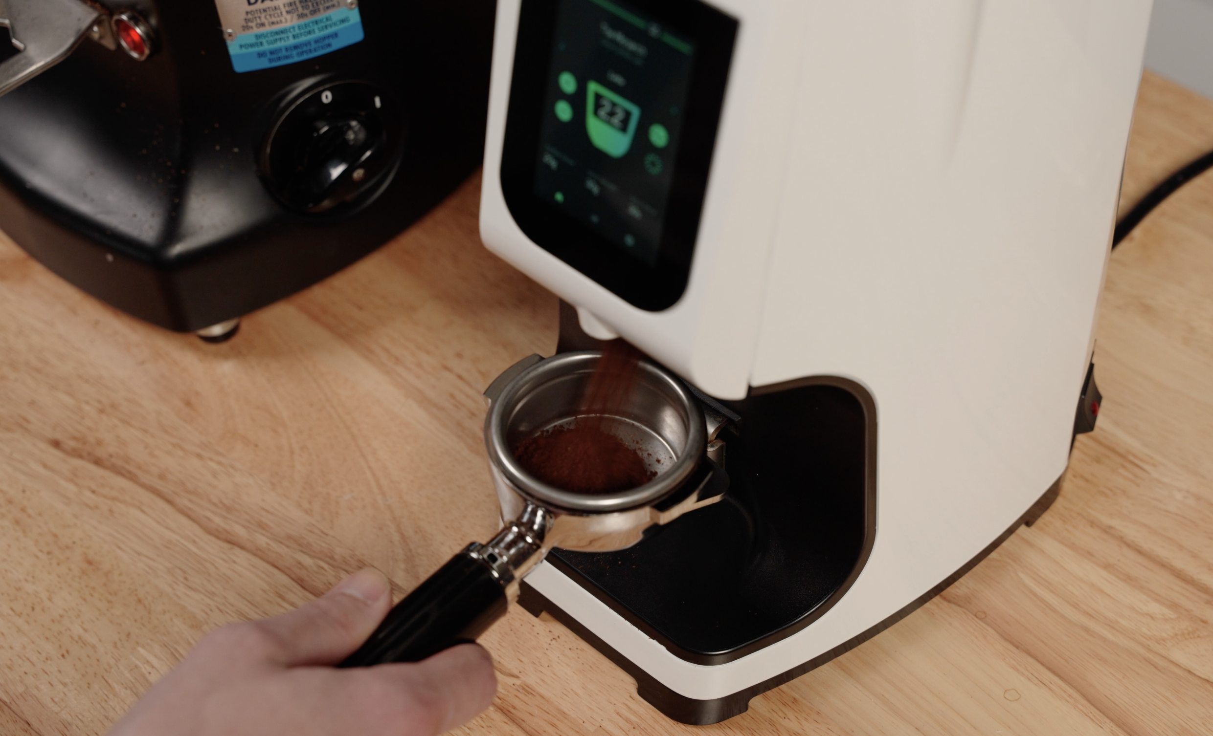Close-up of a person using a coffee grinder with a portafilter underneath, ground coffee is being dispensed into the portafilter, a digital coffee machine is in the background.