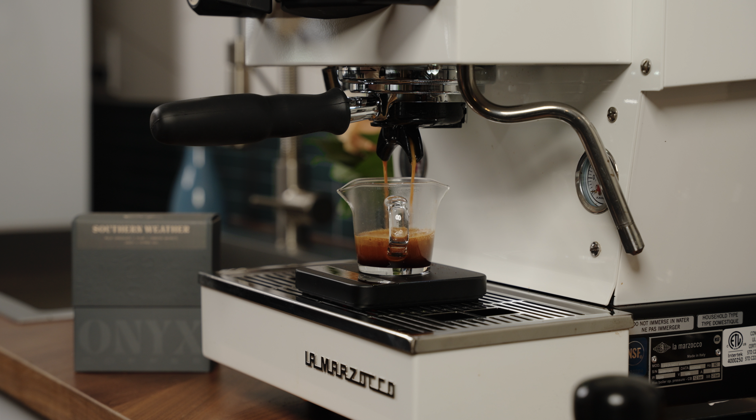 An espresso machine pouring freshly brewed espresso into a glass cup on a digital scale, with a box labeled 'Southern Weather' next to it.