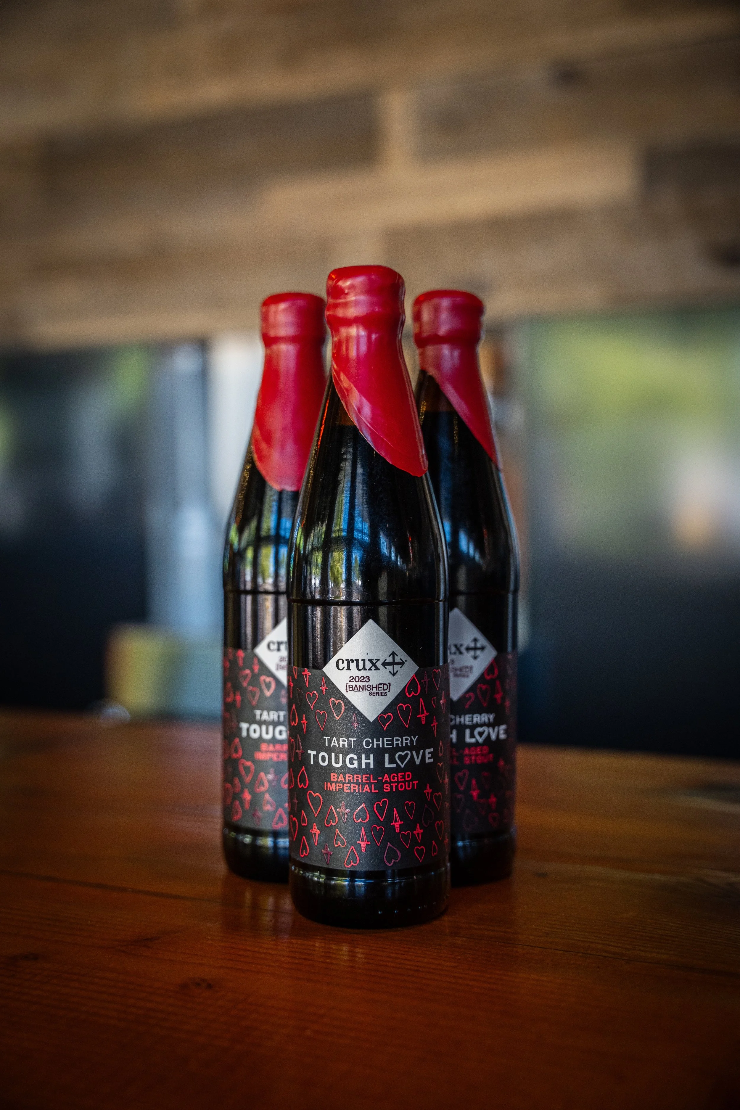Three bottles of tart cherry stout beer with red wax seals on dark glass bottles, placed on a wooden surface.