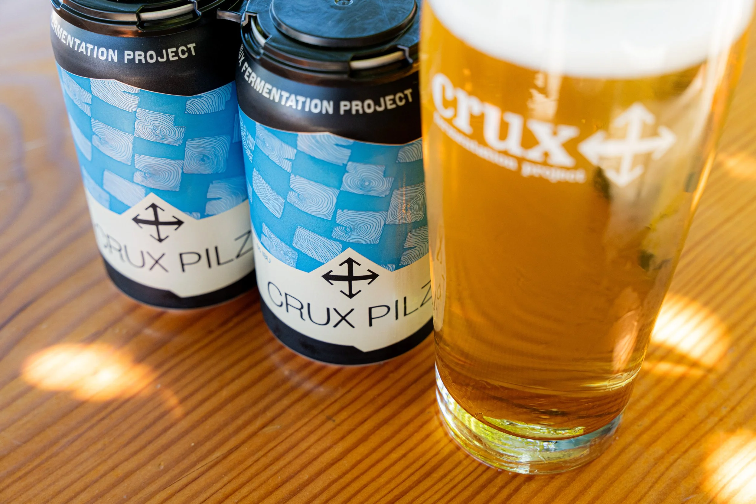 Two cans of Crux Pilz beer on a wooden table alongside a glass of beer with foam on top.