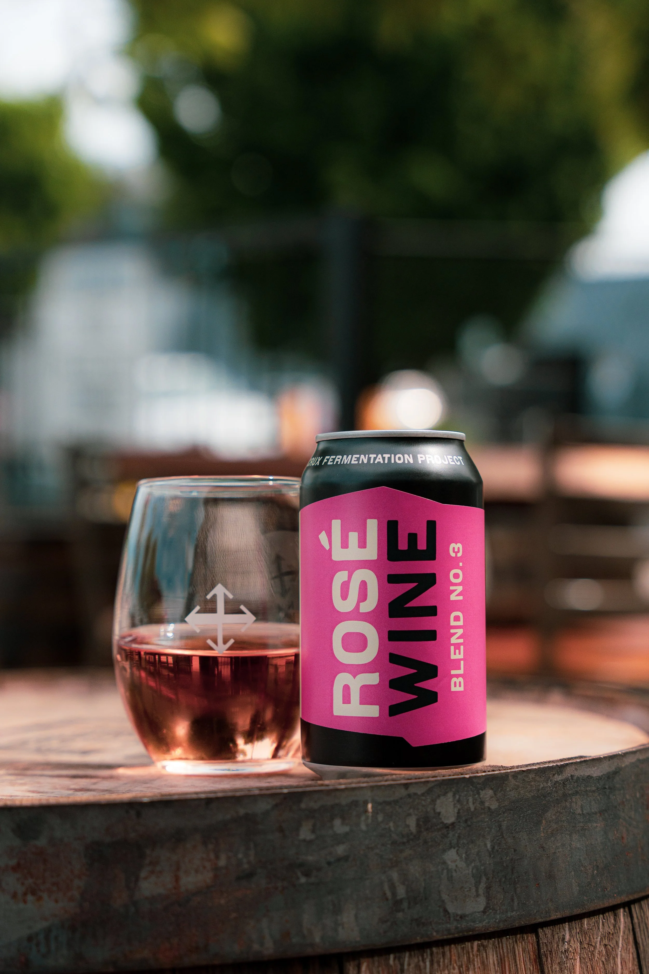 A can of Rose Wine Blend No. 3 stands on a wooden surface next to a glass of rosé wine, outdoors with blurred greenery in the background.