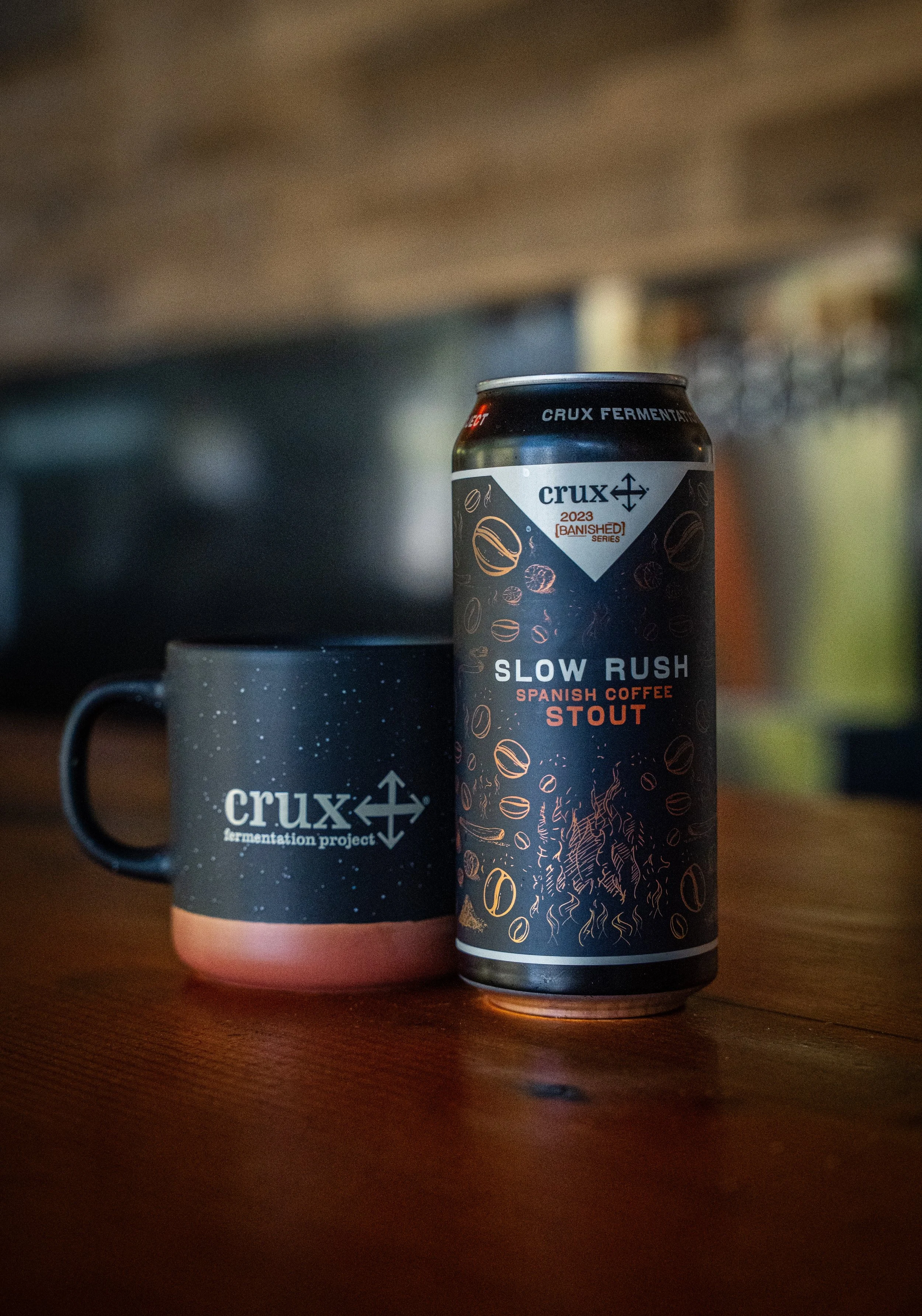 A can of Crux Slow Rush Spanish Coffee Stout beer and a Crux armamentation project mug on a wooden surface.