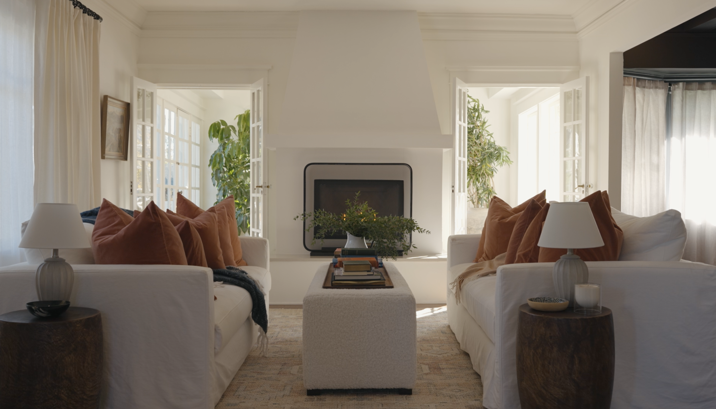 Living room with white sofas, brown pillows, wooden side tables, lamps, a fireplace, and large windows with curtains