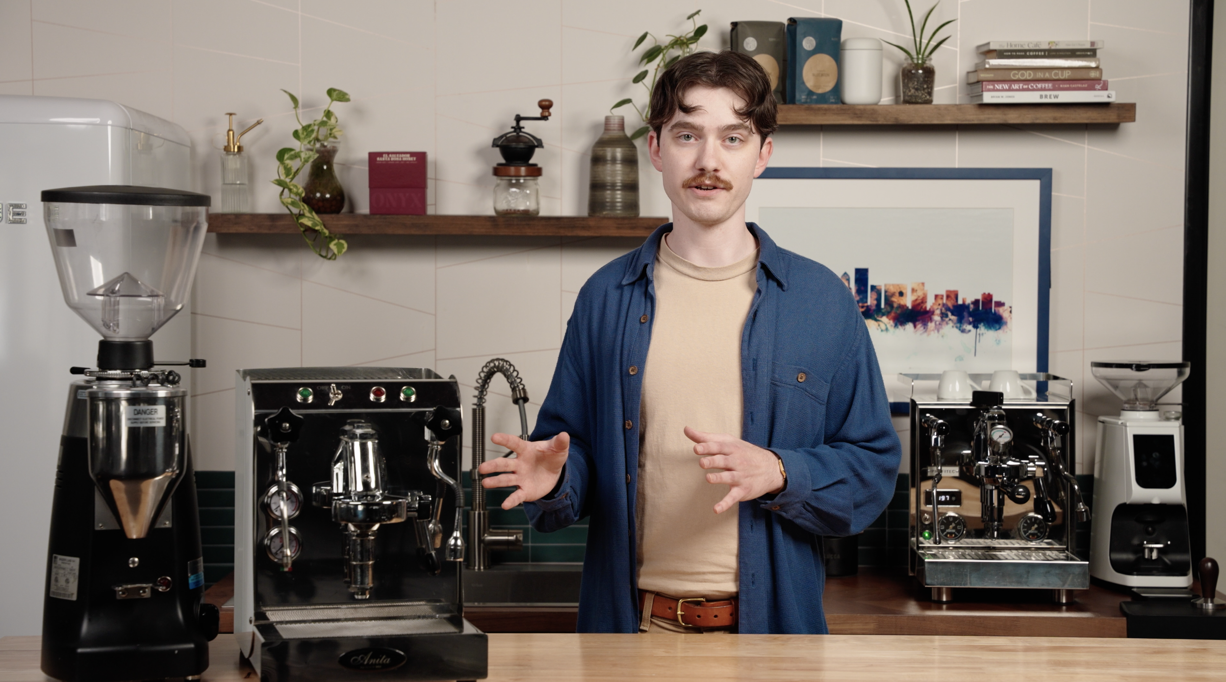 A young man with dark hair, a mustache, wearing a beige t-shirt and a blue shirt, standing behind a wooden counter in a coffee shop or cafe, surrounded by coffee-making equipment, including a coffee grinder, espresso machine, and a scale, with shelve