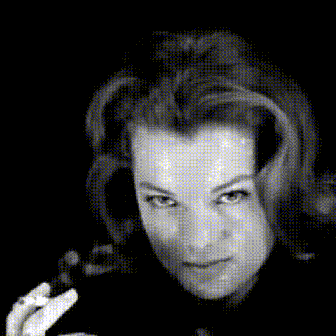Black and white close-up of a woman with styled hair, looking directly at the camera with a confident expression and holding a cigarette.