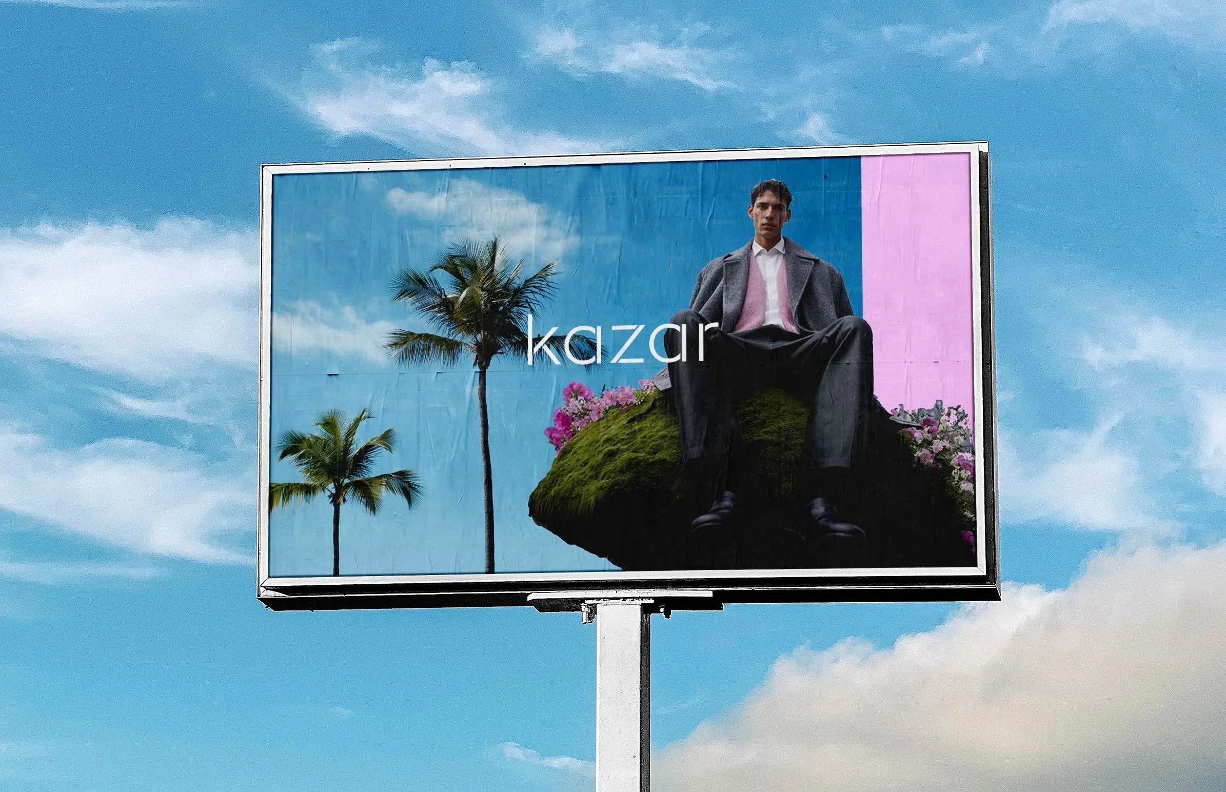 Billboard advertisement with a young man sitting with crossed legs, wearing a gray coat and pink sweater, against a backdrop of palm trees and a sky reflected in glass. The billboard has the word "kazar" in white text on the left side.