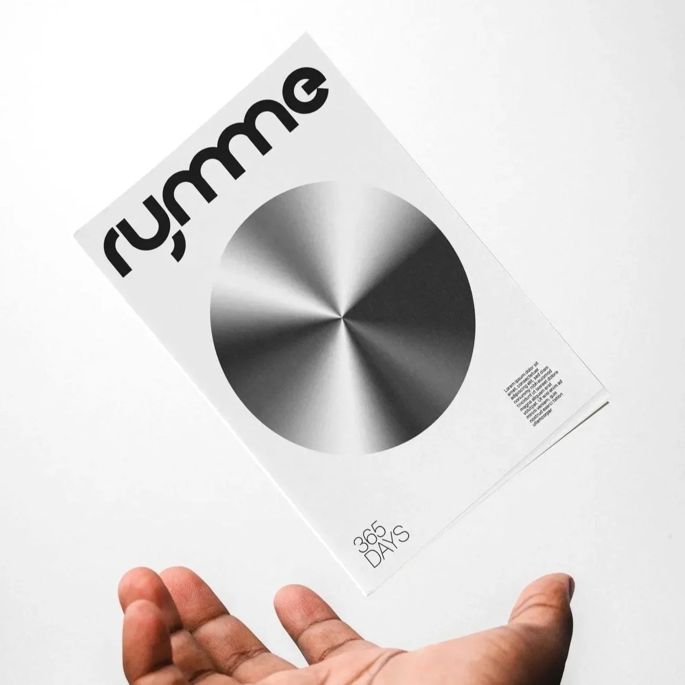A hand holding a magazine with a black and gray circular pattern and the word 'rumme' in bold black letters at the top. The magazine also features the text '365 DAYS' at the bottom.