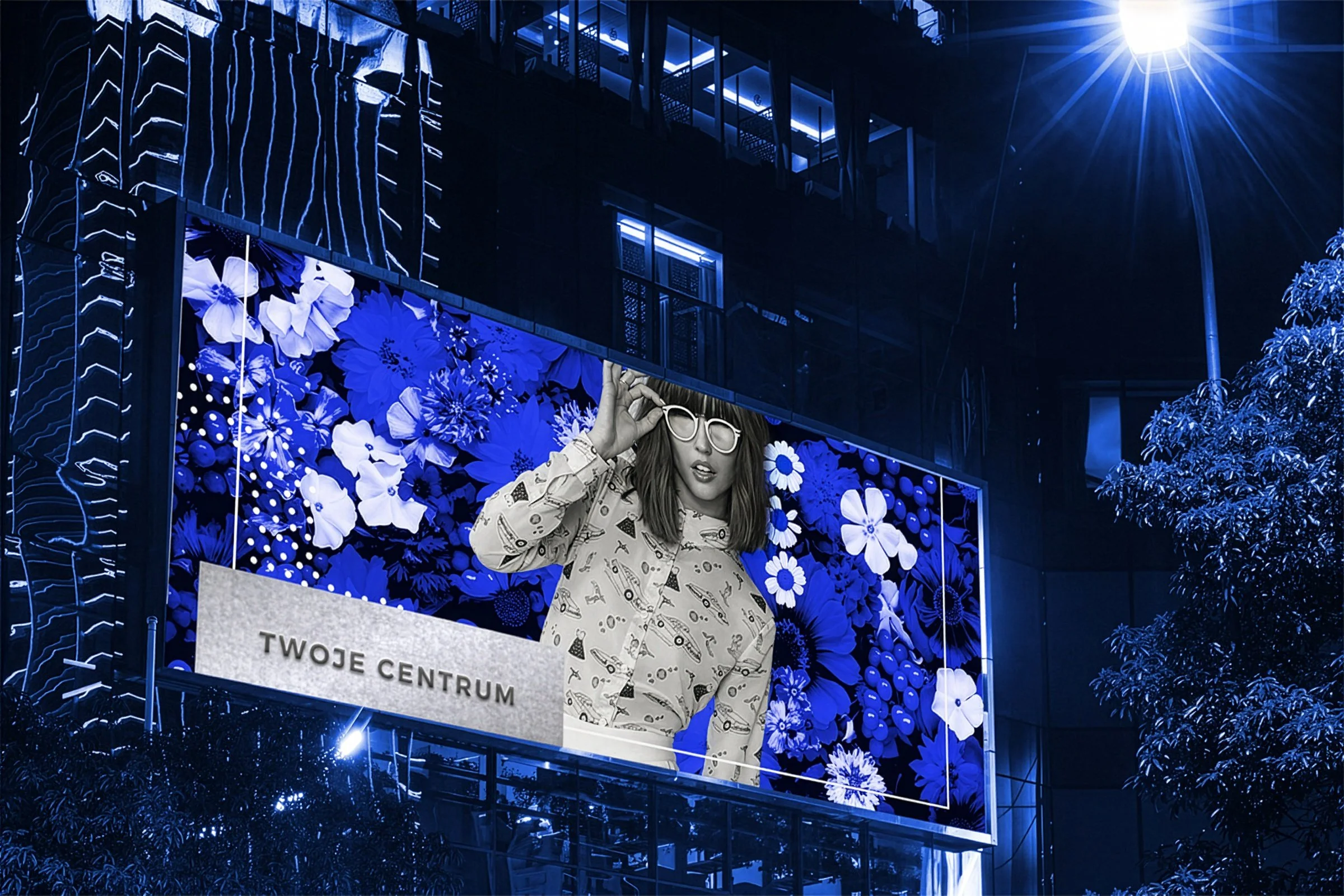 Nighttime city scene with bright streetlights, a colorful digital billboard featuring a woman with glasses against a blue floral background, and trees illuminated by artificial lights.