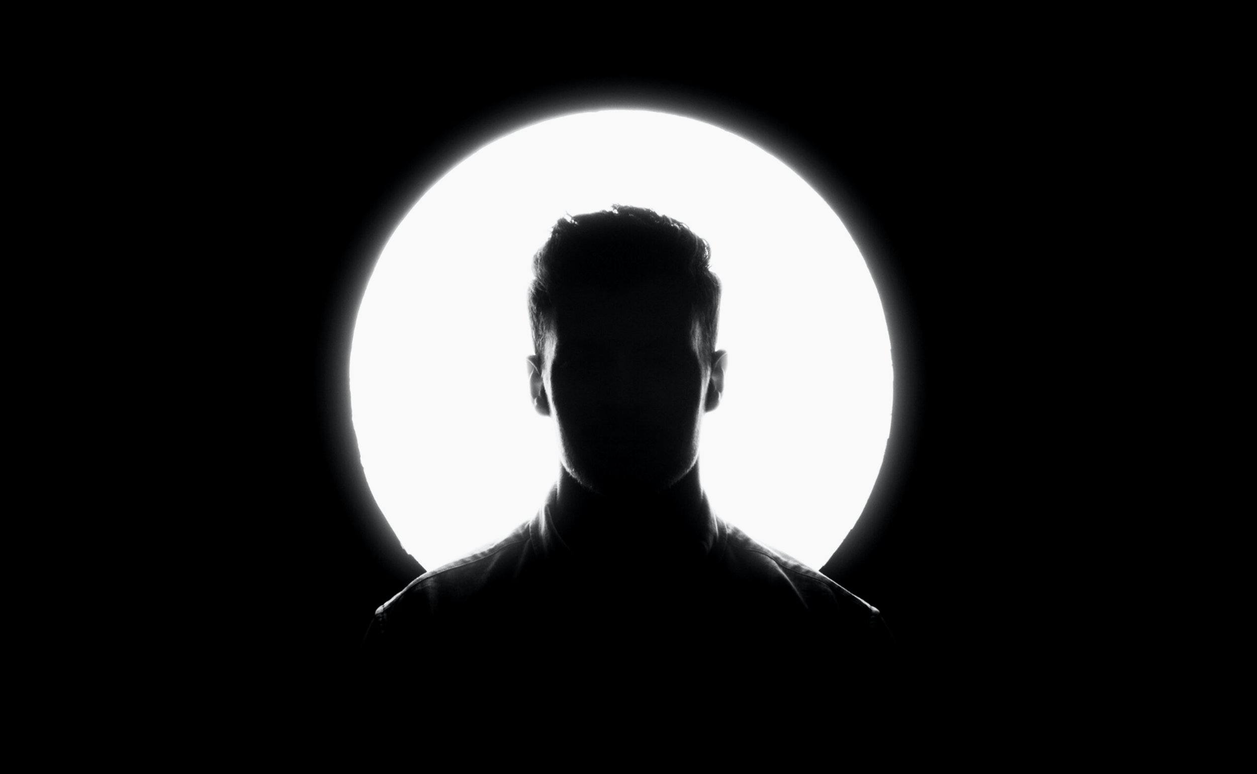 A black silhouette of a person facing forward, with a bright white circular light behind their head, creating a halo effect, against a black background.