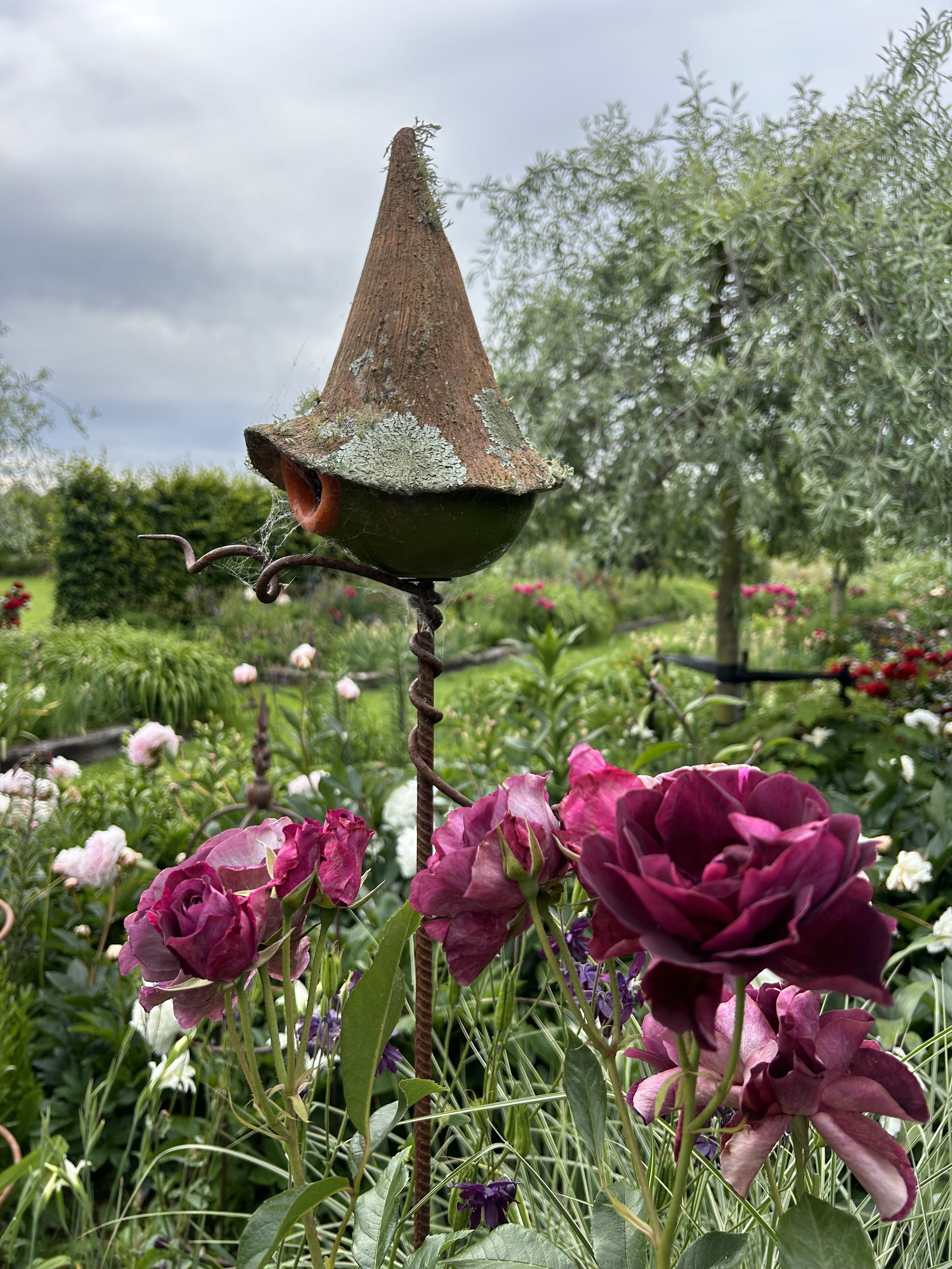 Quirky birdhouse surrounded by burgundy Iceberg roses and pink Sarah Bernhardt peonies in the background.  The tree is a weeping ornamental silver pear tree.  