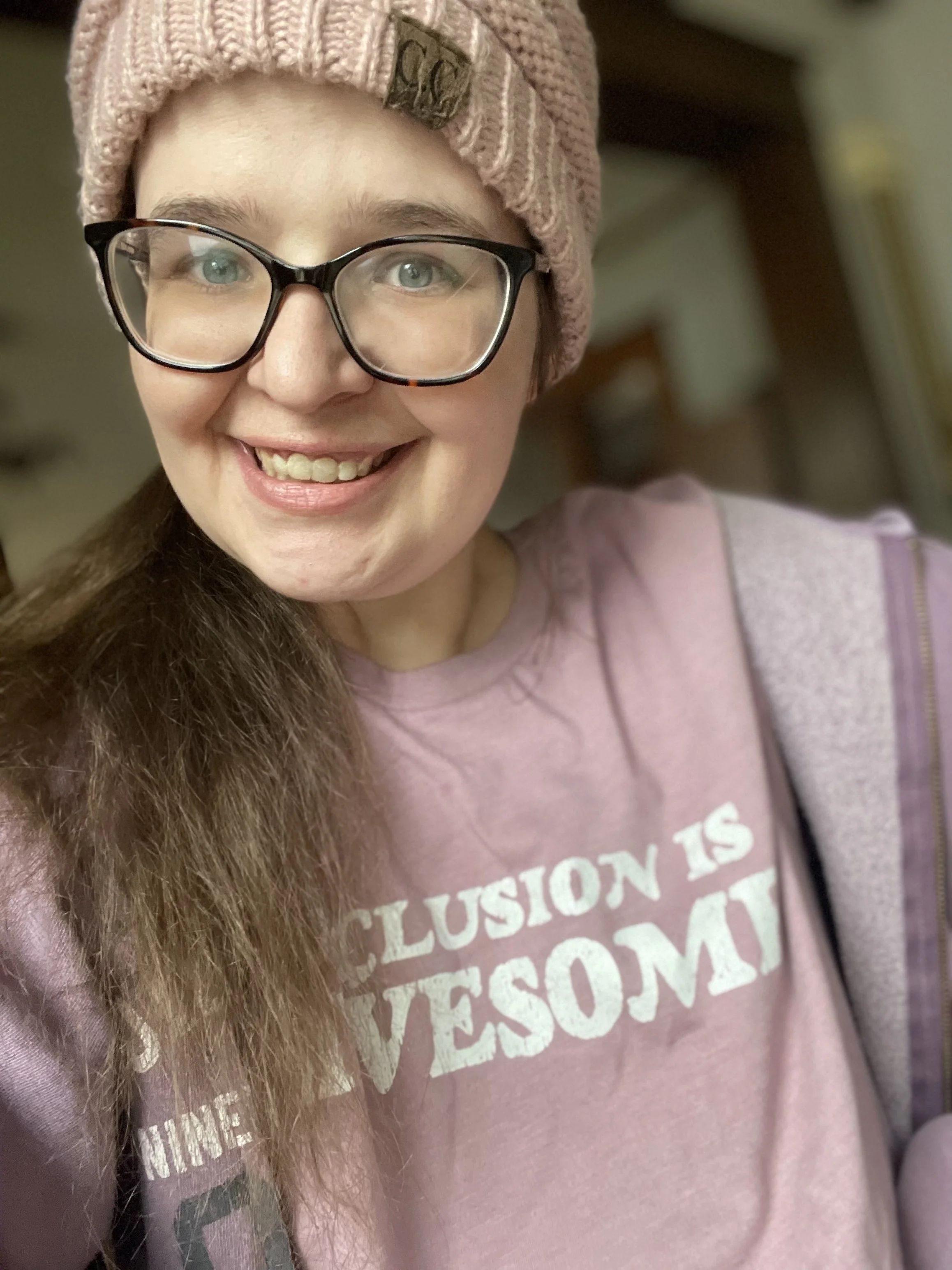 Annora Ruth wearing a pink beanie, pink shirt about inclusion. She is wearing glasses and has brown hair in a ponytail
