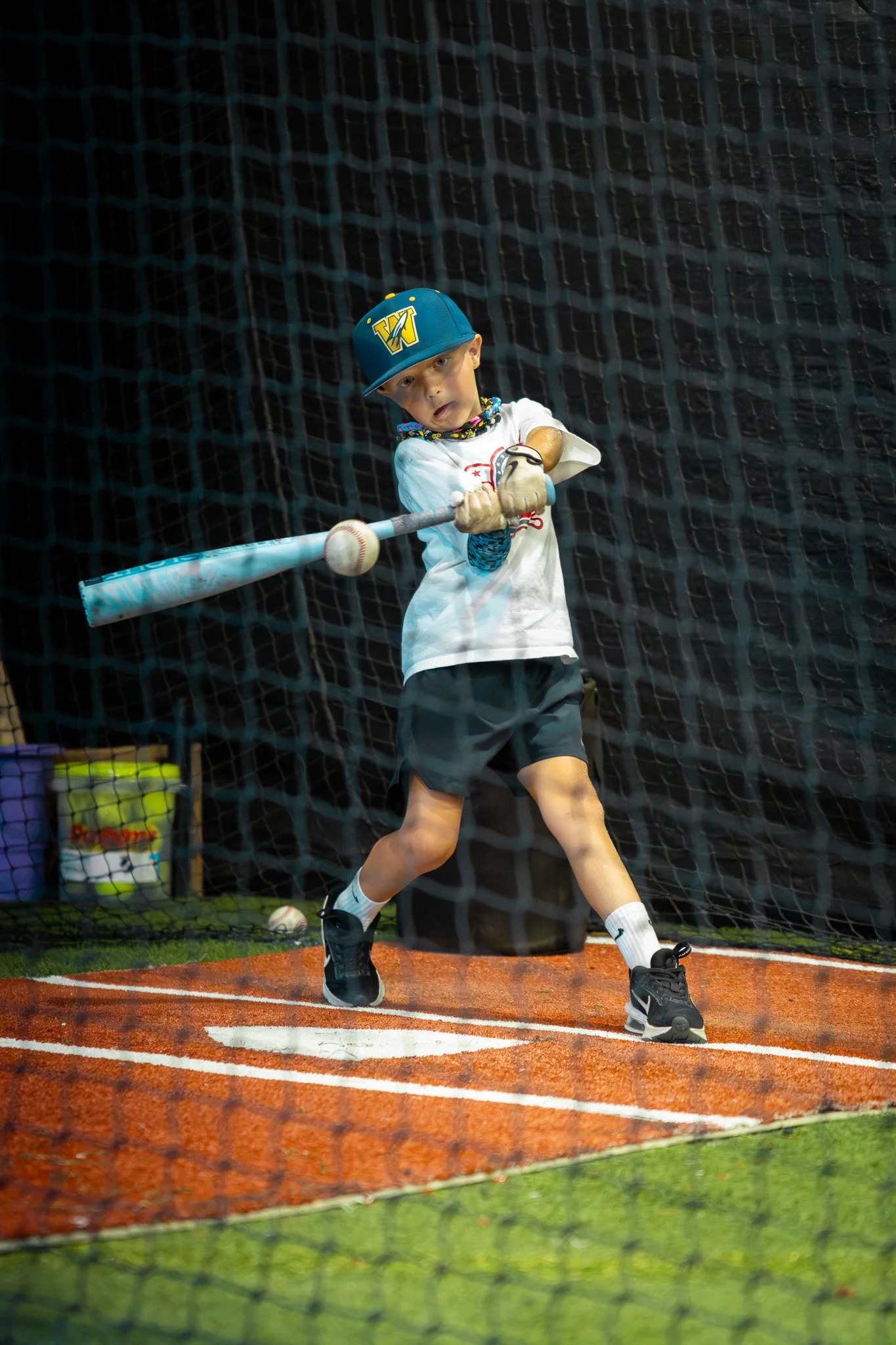 A young boy practicing baseball hitting in an indoor batting cage, swinging a bat at a ball.