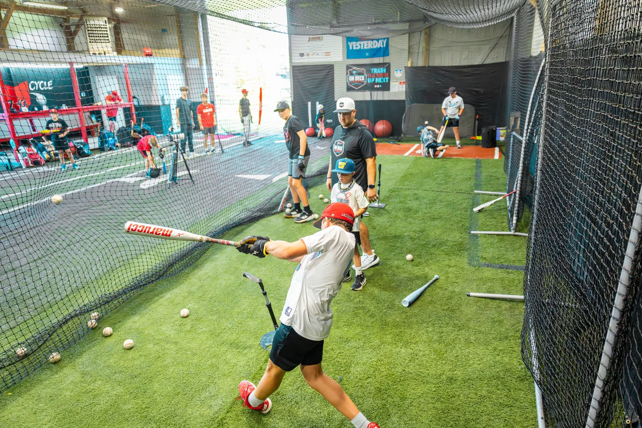 A young boy in a white shirt with a red cap swings a baseball bat at a batting cage at an indoor batting facility, with other children and adults observing and participating in baseball practice.