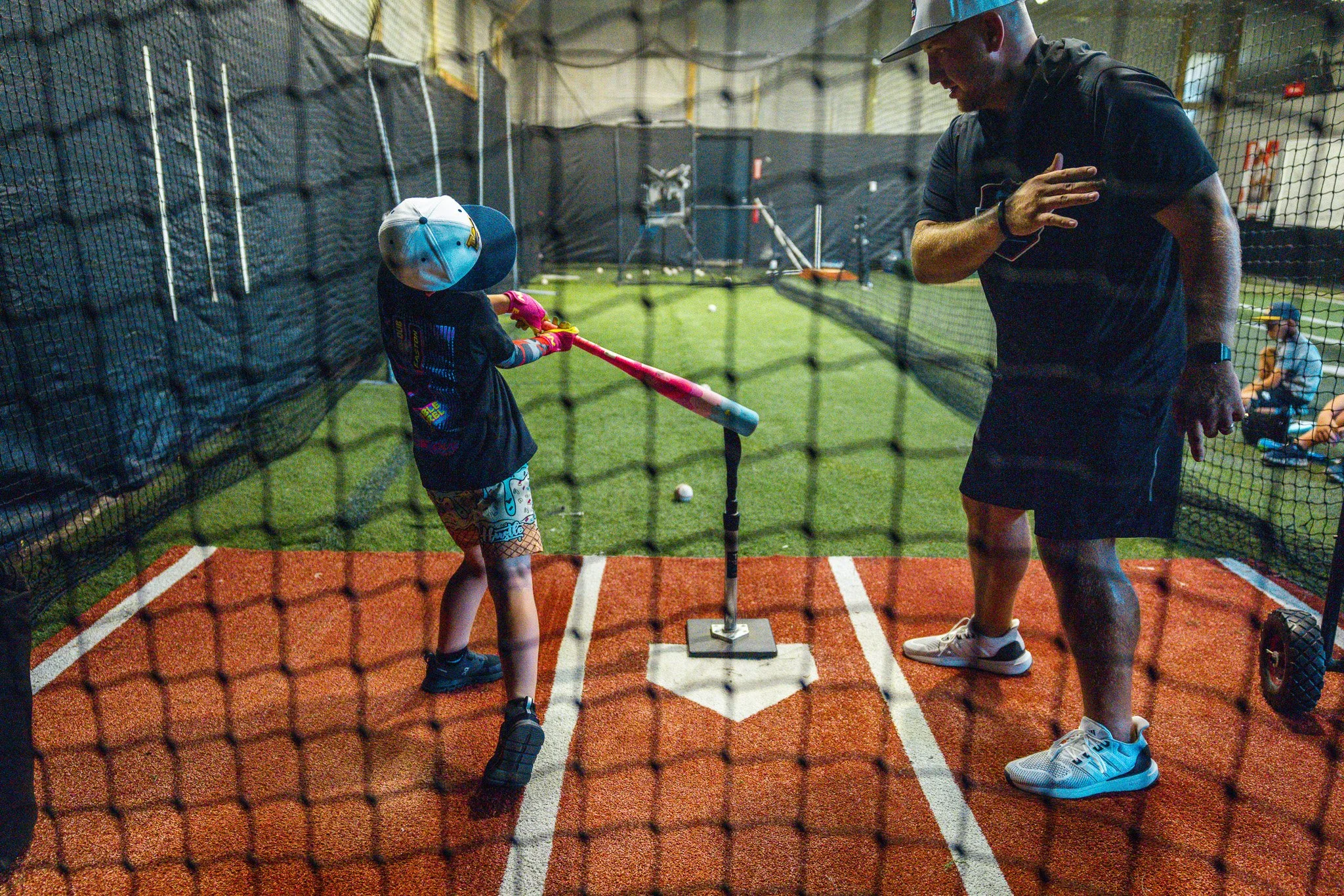 A young child practicing baseball in an indoor batting cage, swinging a pink bat while an adult coach stands nearby, supervising. The cage has a netting as a barrier, and the floor is designed for baseball practice.