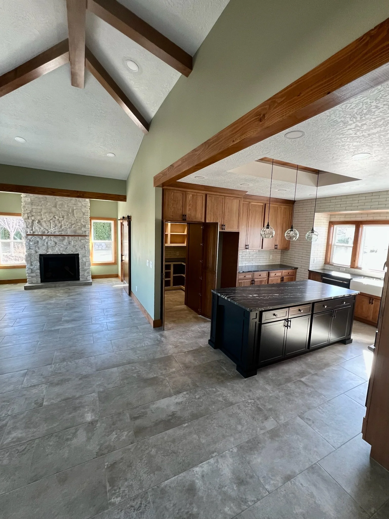 Open-concept kitchen and living room with stone fireplace, large windows, wooden cabinets, black kitchen island, pendant lighting, and tiled flooring.