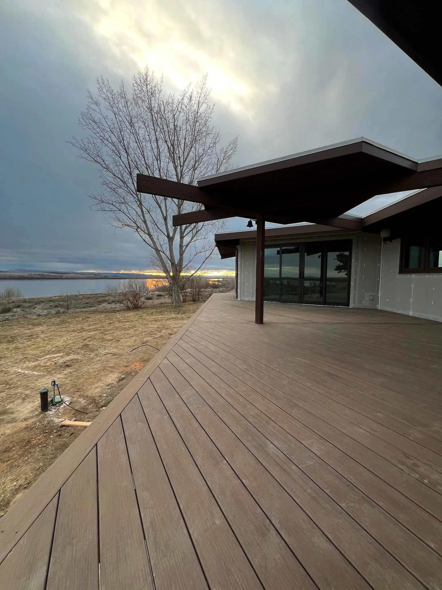 Partial view of a wooden deck extending from a house, with a large leafless tree and body of water in the background under a cloudy sky at sunset.