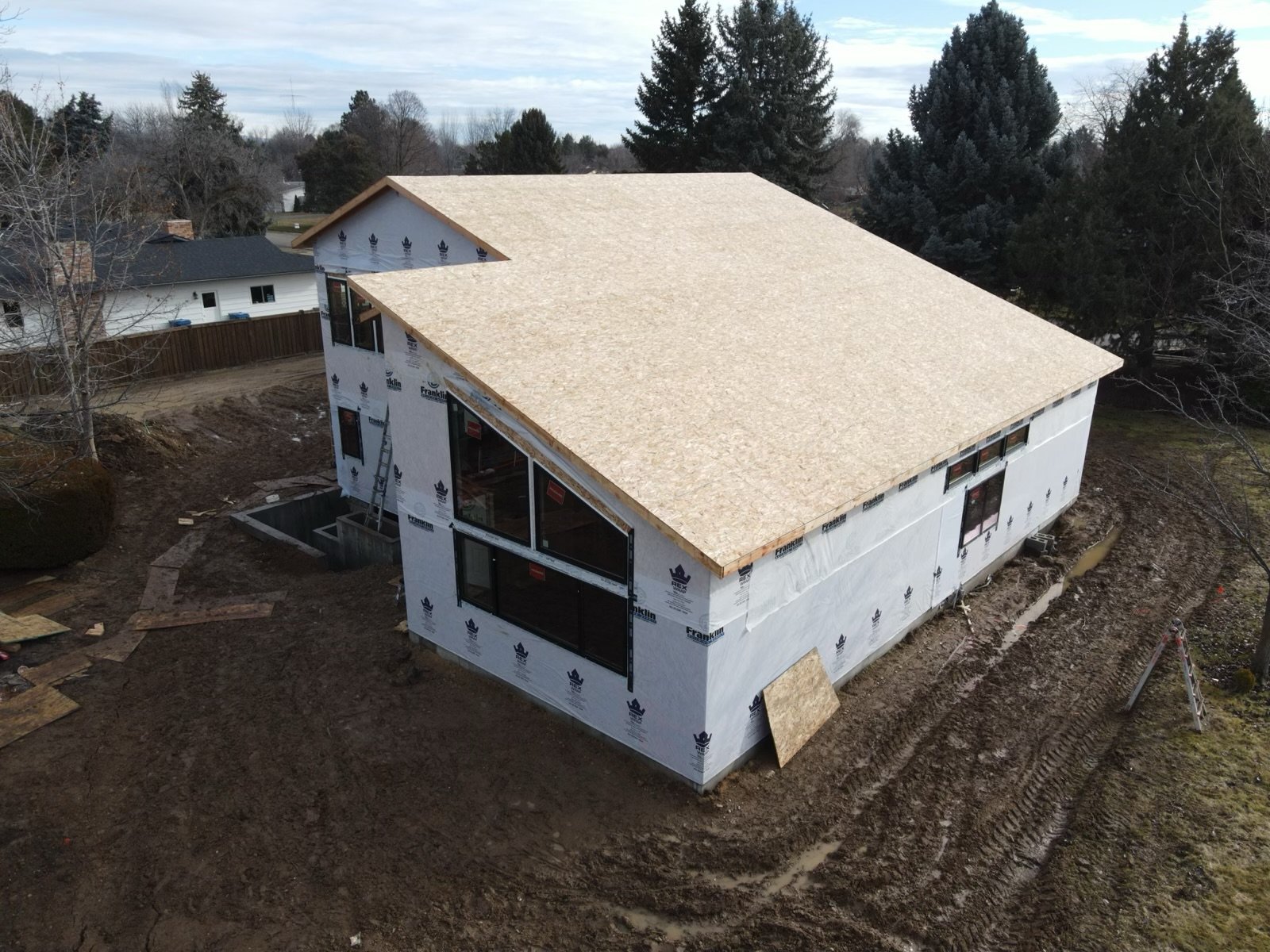 A house under construction with a partially finished roof and exterior walls covered in weather barrier sheathing. The house is on a dirt lot with construction materials and a ladder nearby, surrounded by trees and neighboring houses.