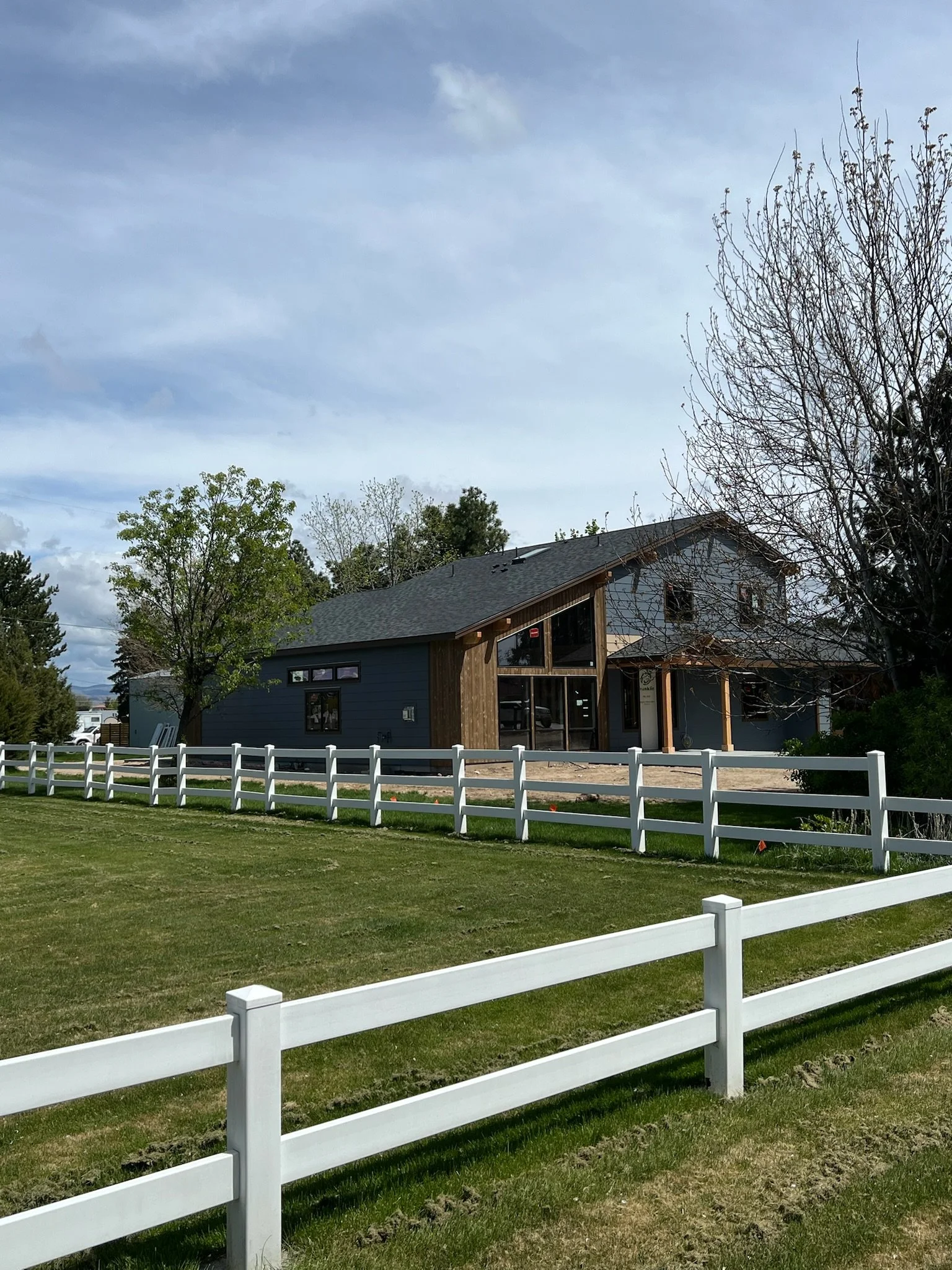 A modern house with large glass windows and a wooden accent wall, surrounded by a green lawn and a white fence, with trees and a blue sky in the background.