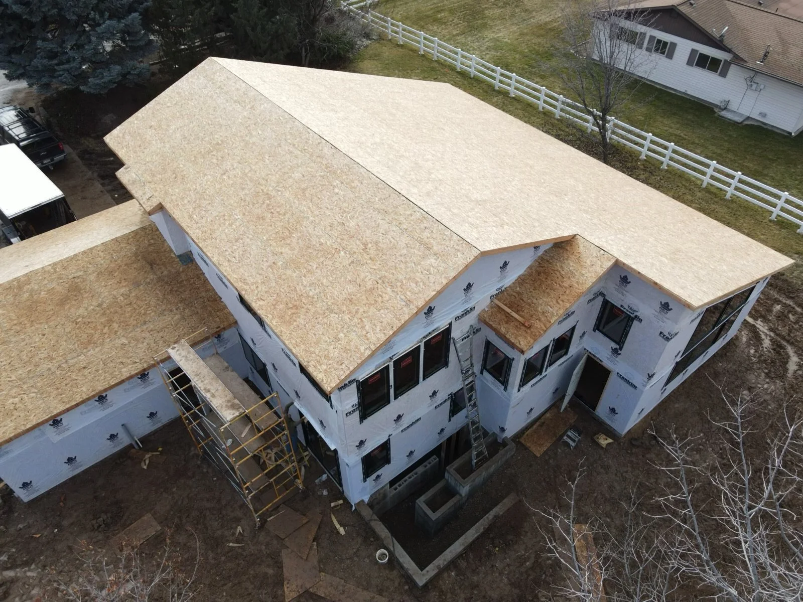 An aerial view of a house under construction with new wooden roof sheathing and wrapped exterior walls, surrounded by a dirt yard and neighboring houses.