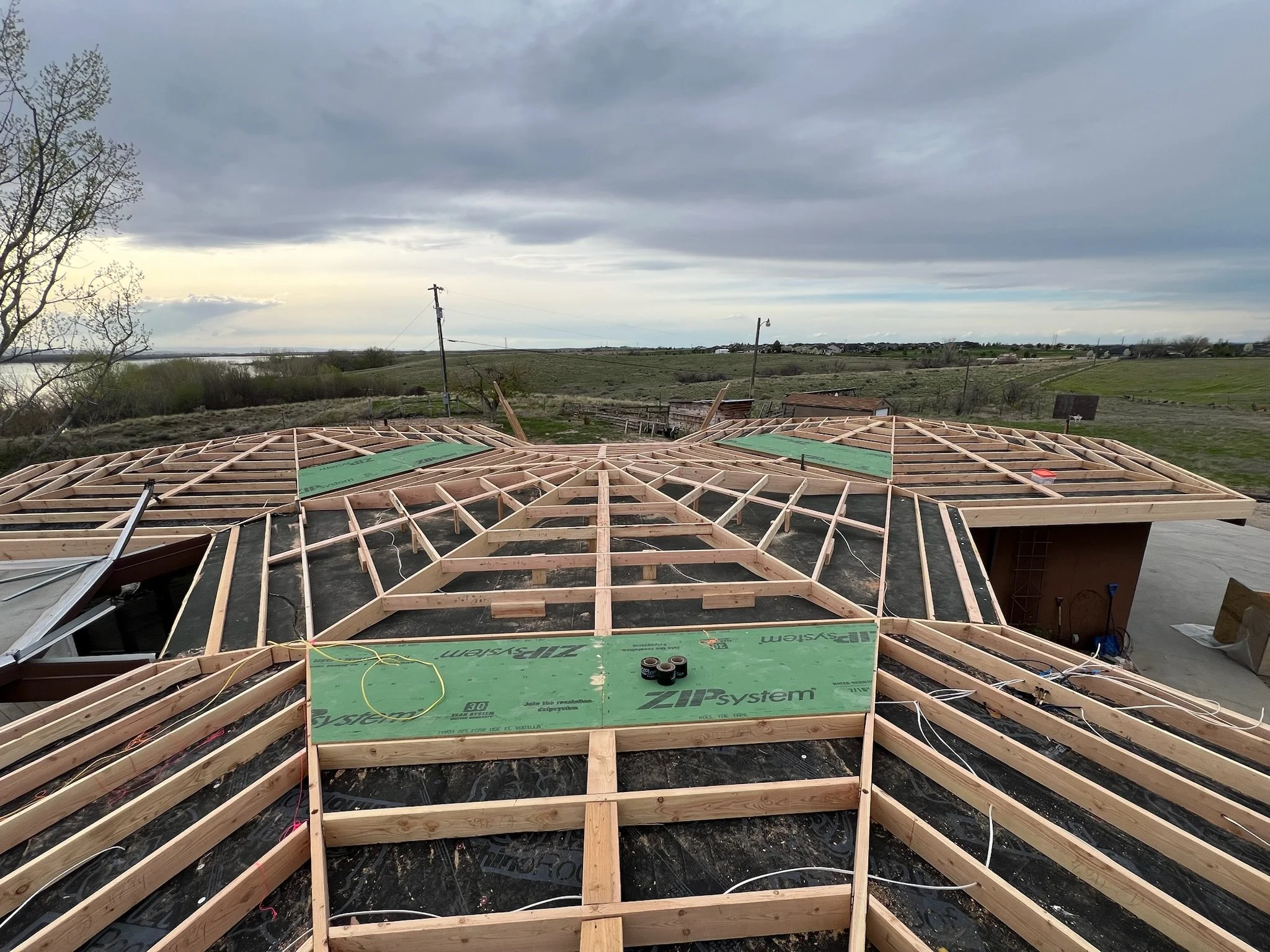 Construction site with wooden roof framing on a house, overlooking a rural landscape with power lines and cloudy sky.
