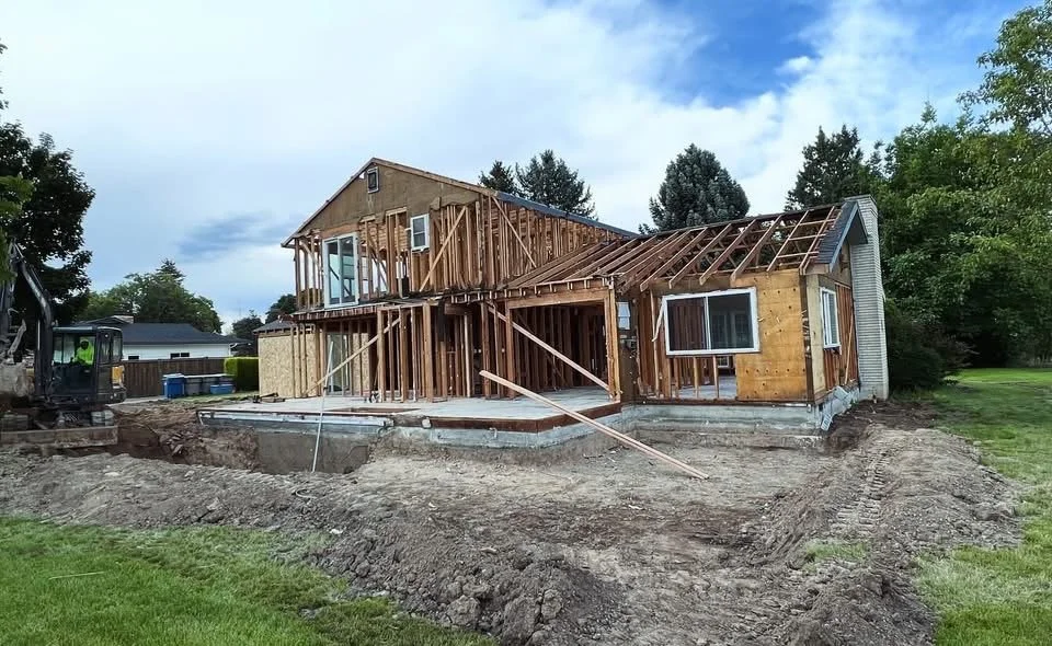 House under construction with exposed wooden framing, on a cleared dirt lot with construction equipment nearby.