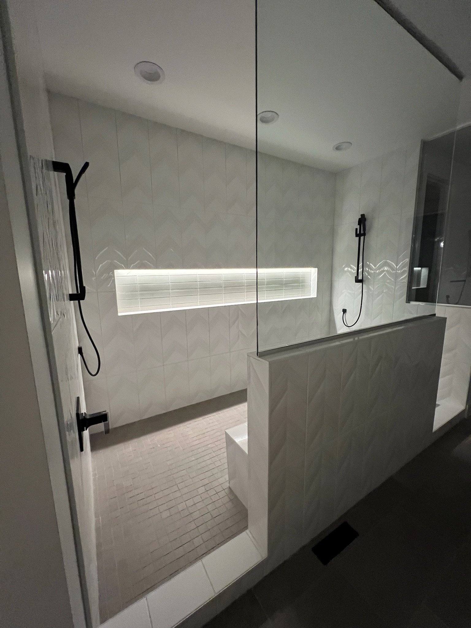 Modern walk-in shower with white tiled walls, two black adjustable showerheads, and a built-in bench, separated by a half-wall, with a backlit horizontal niche.