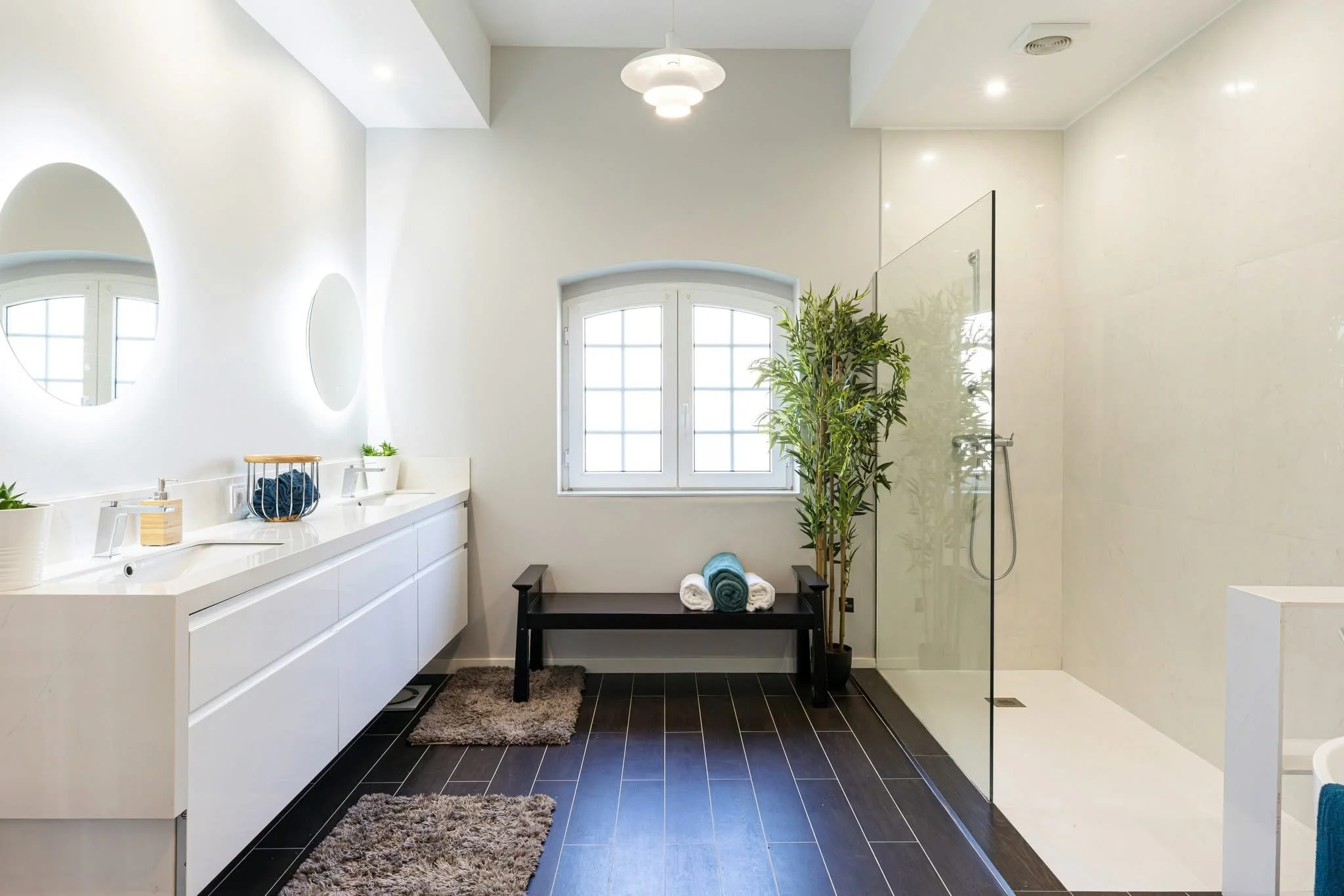 Modern bathroom with double sinks, large mirrors, a shower with glass door, dark wood flooring, and a window with white frame. There are towels, a bench, and indoor plants.