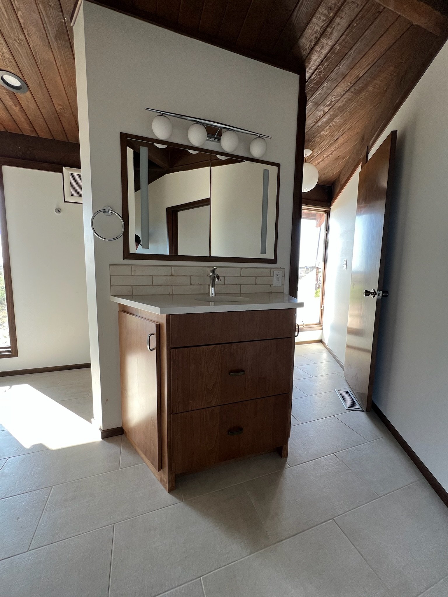 A small wooden vanity with a white countertop and a sink, located in a room with tiled flooring and a mirrored wall above it. There is a four-bulb light fixture above the mirror. Natural light is coming from a window or door to the right.