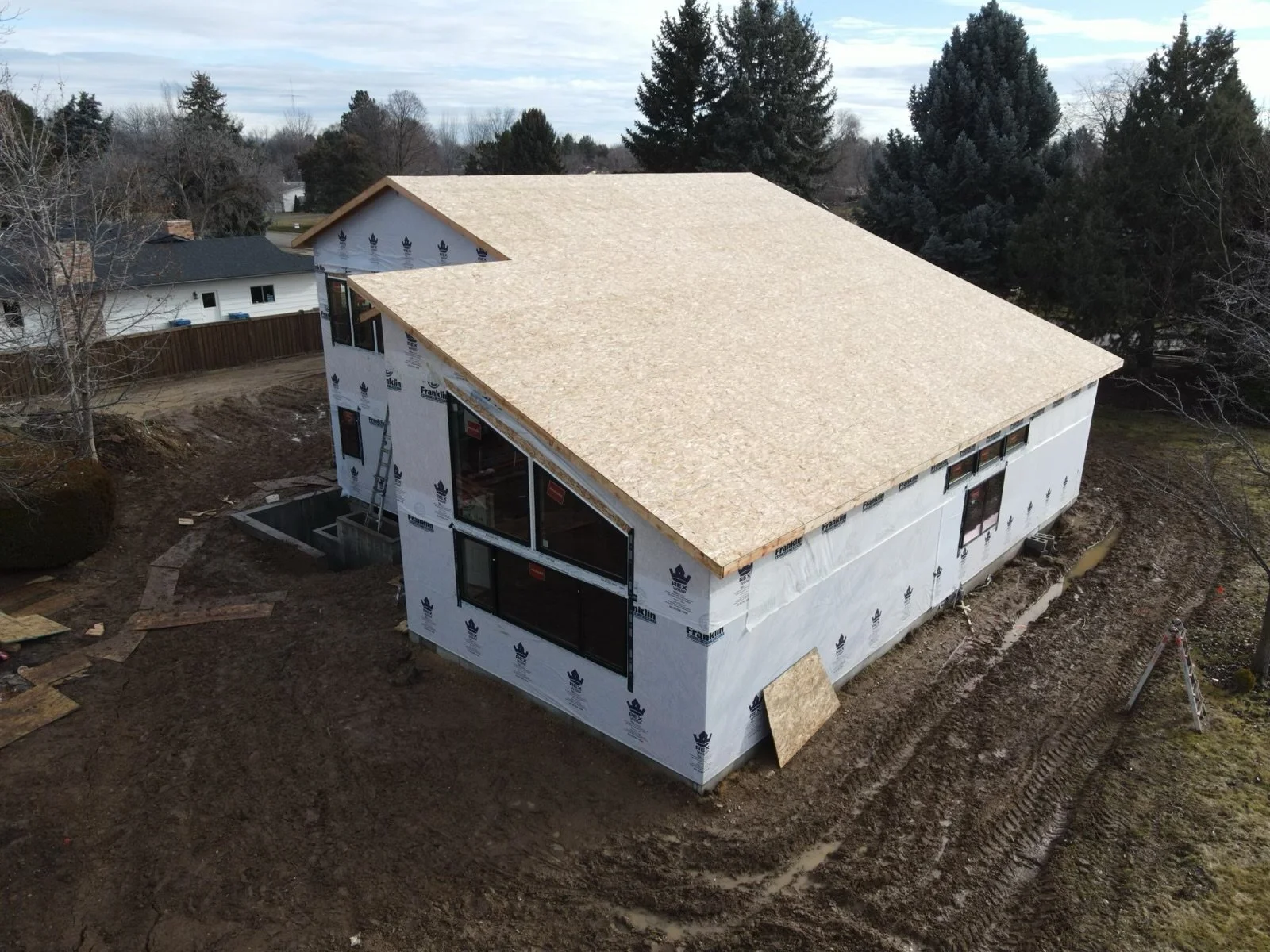 A house under construction with a new roof and exterior wrap, surrounded by dirt and construction materials, with trees and neighboring houses in the background.