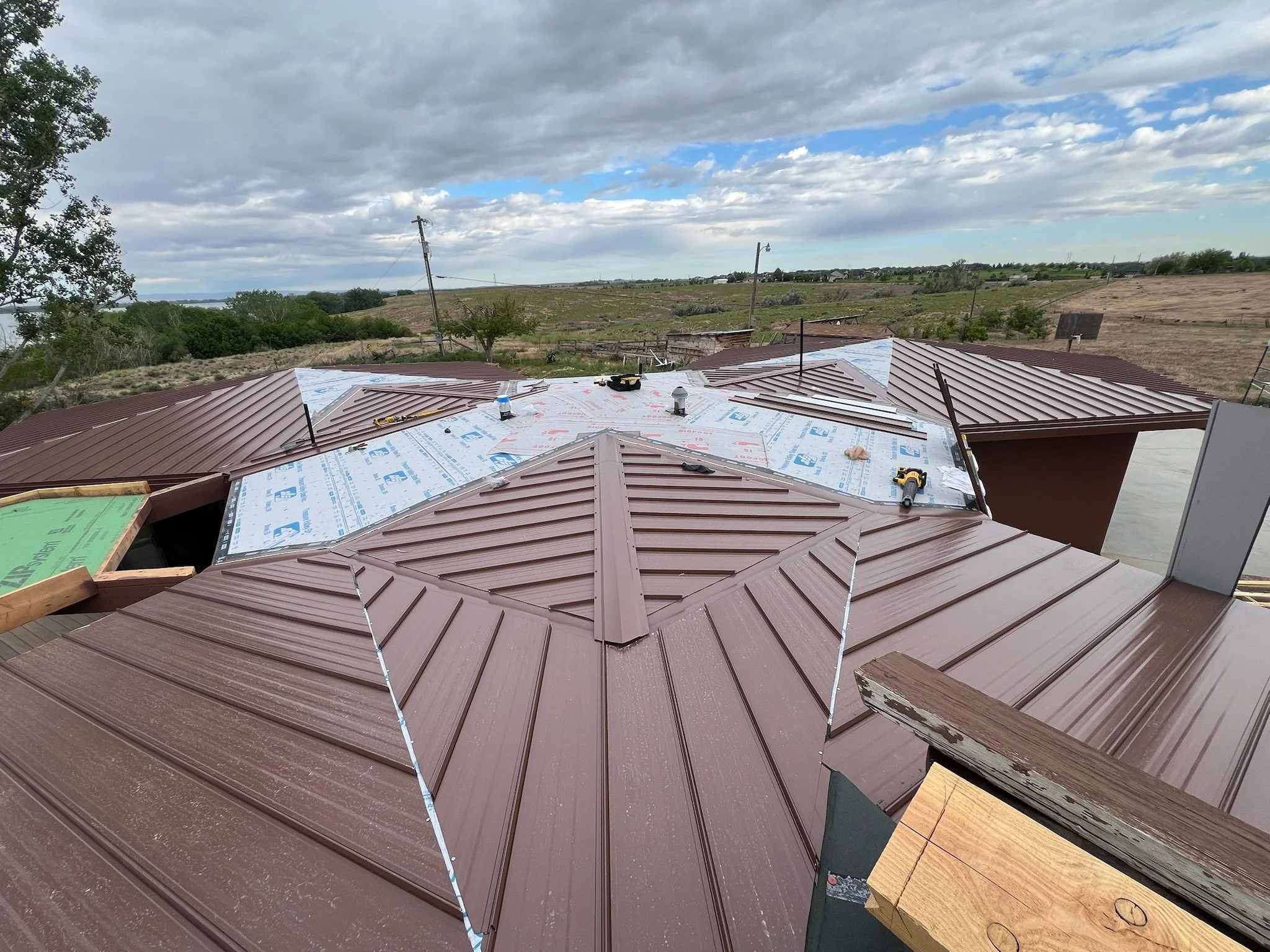 Construction site on a rooftop with partially installed brown metal roofing, tools, and materials, under a cloudy sky with open landscape in the background.