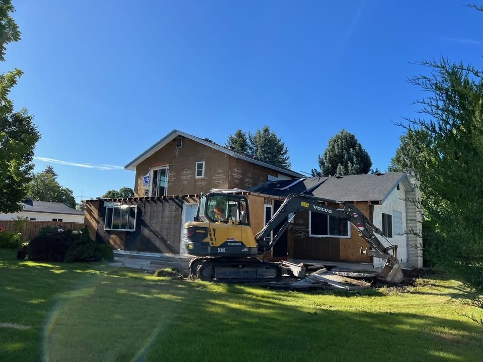 Construction work on a house with a mini excavator demolishing a section of the exterior wall on a bright, sunny day.