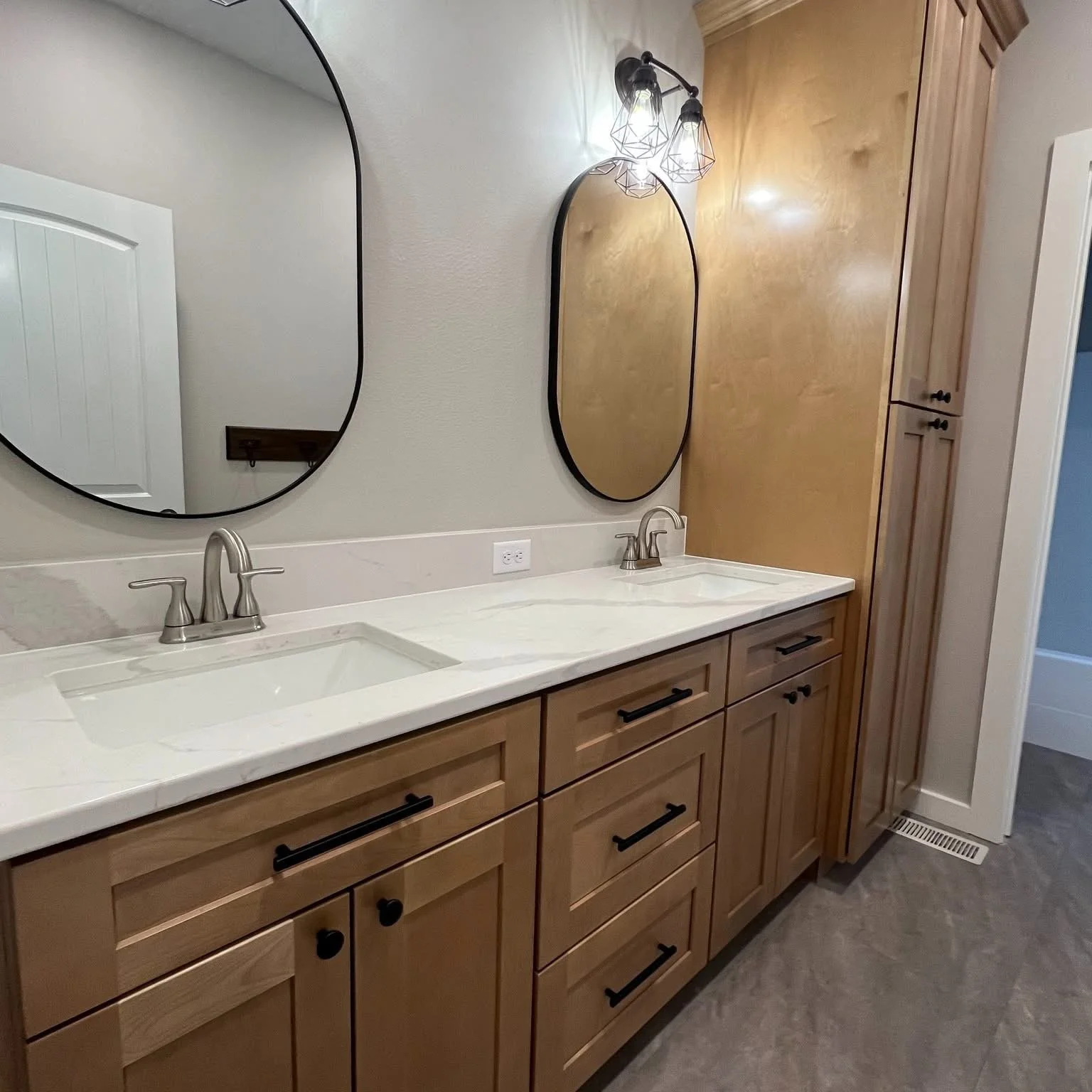 Double bathroom vanity with light wood cabinets, white marble countertops, two oval mirrors with black frames, and modern light fixtures with exposed bulbs.