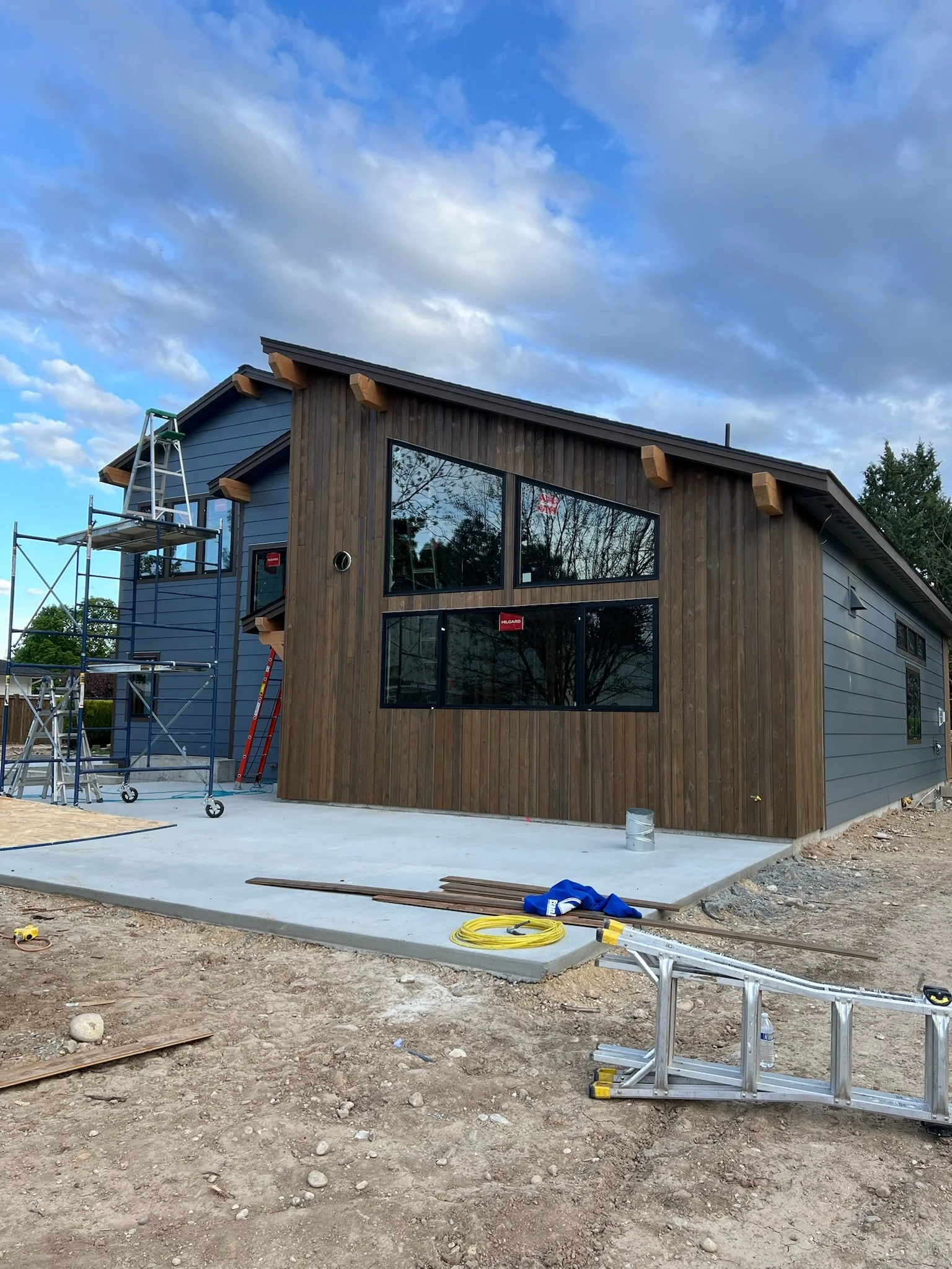 Construction site of a modern house with large windows, partly finished exterior, scaffolding on the side, and construction tools on the ground