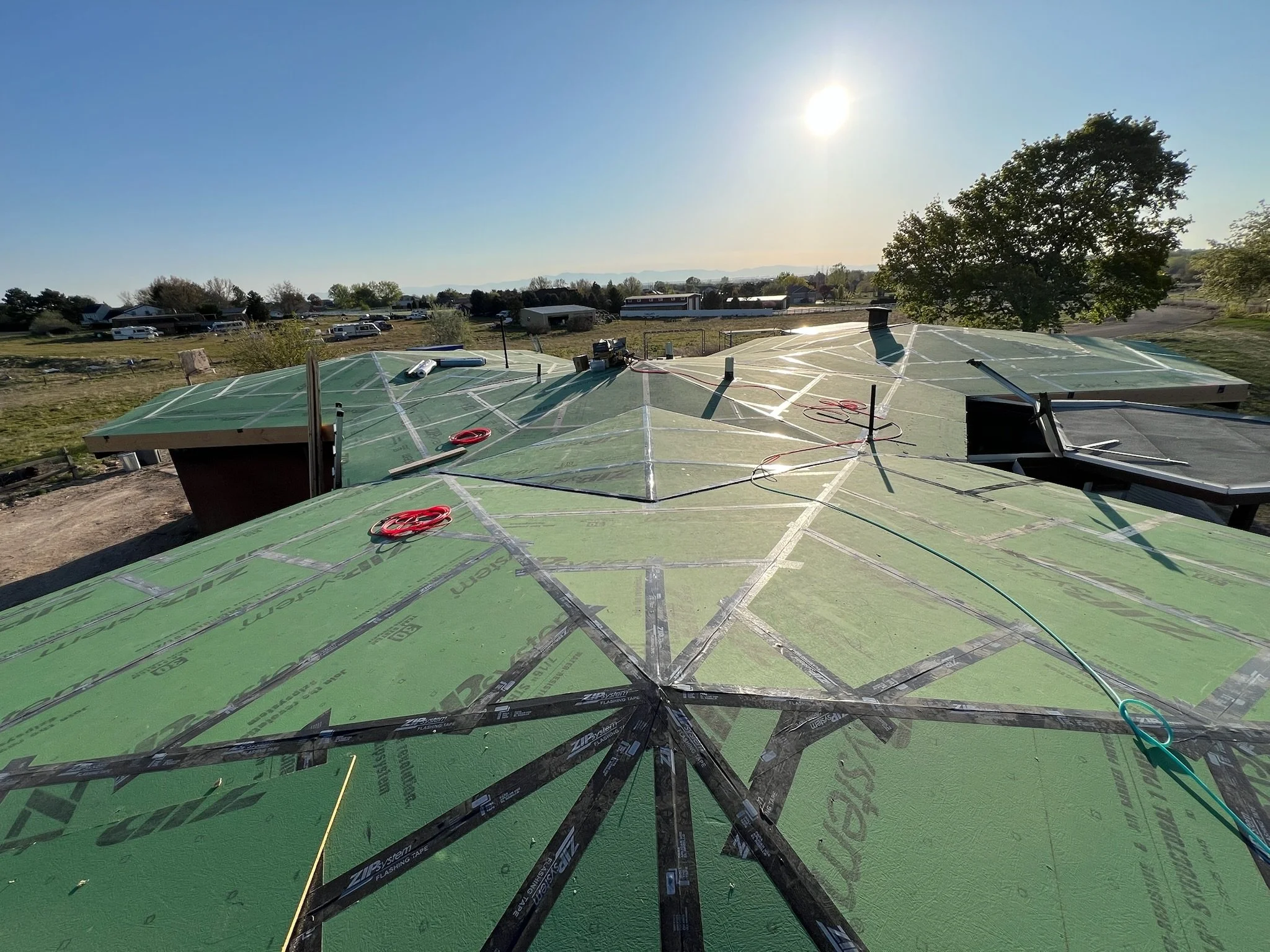 Construction site on a building roof with green sheathing and black tape, tools, and electrical cords visible, under a clear sunny sky with a tree and distant structures in the background.