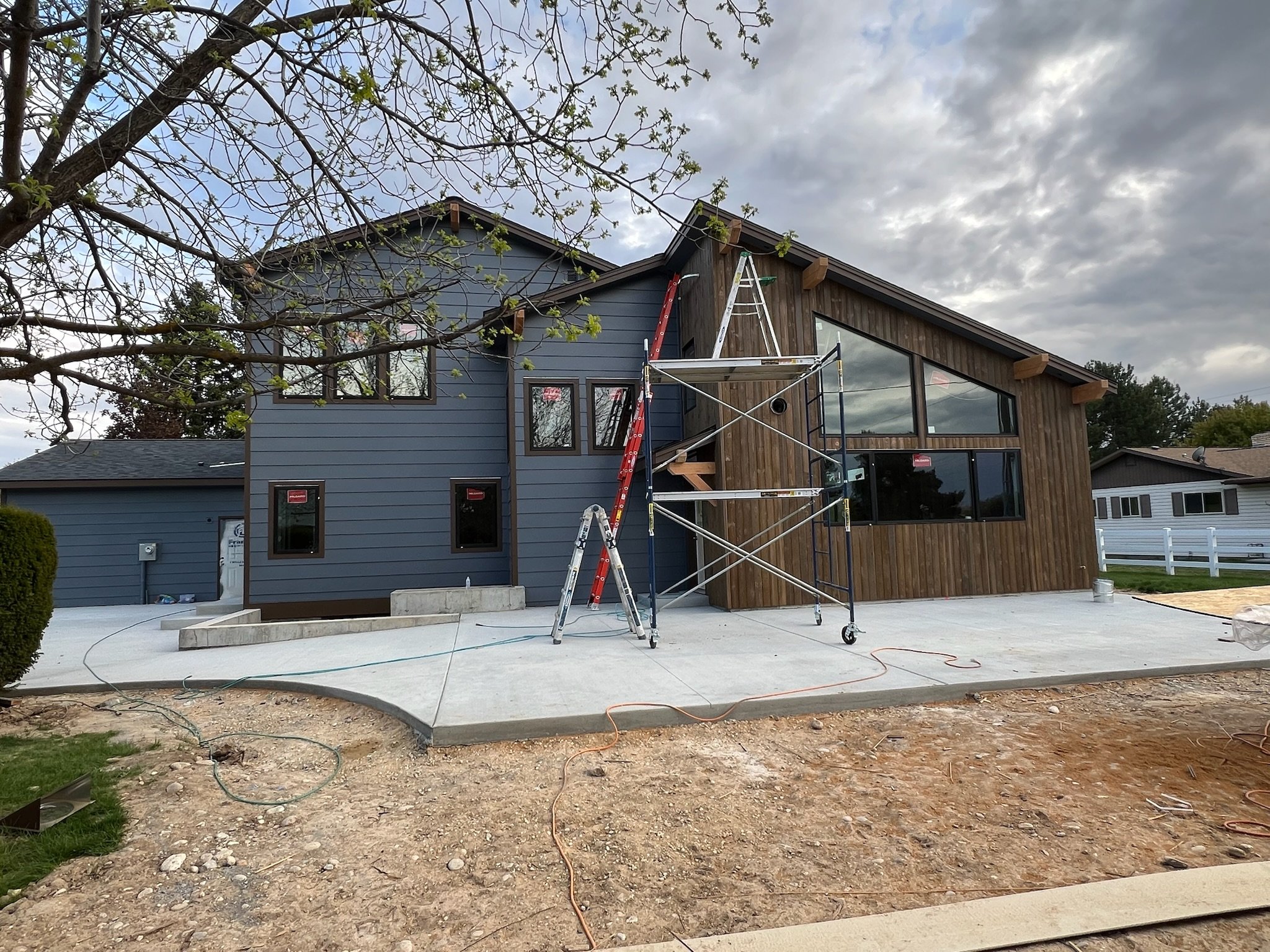 A modern house under construction with scaffolding and ladders in front. The house has blue and wood-paneled siding, large windows, and a concrete patio. Construction tools and materials are visible.
