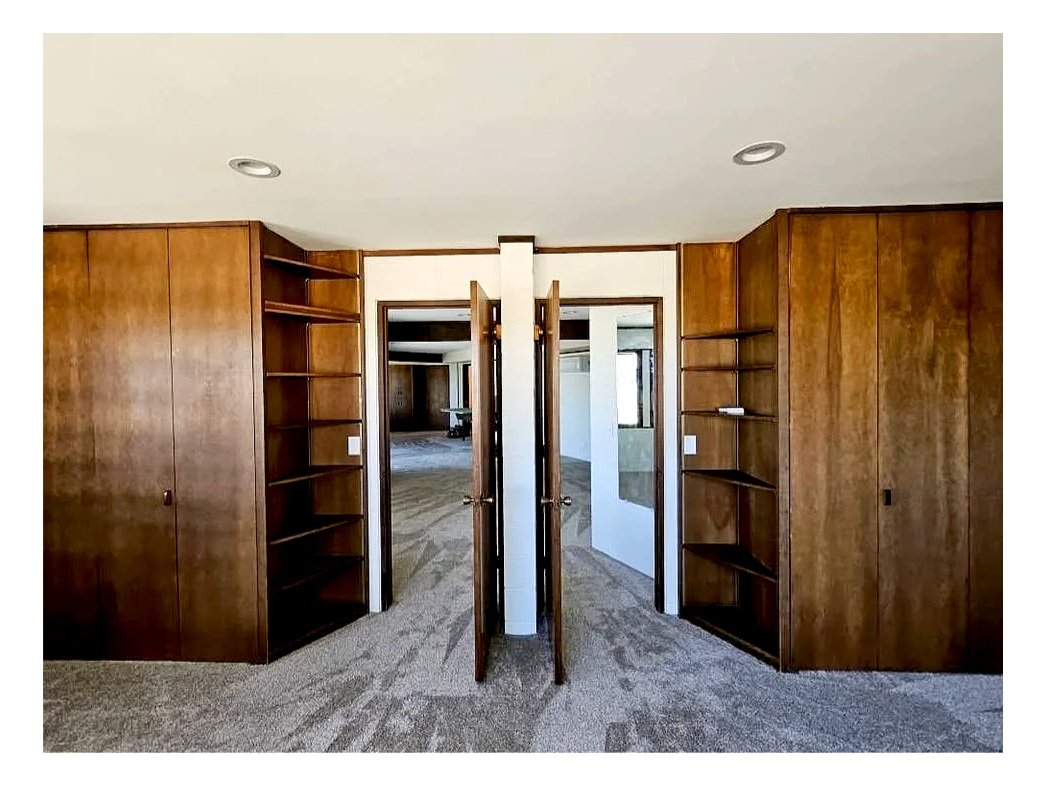 Empty room with wood-paneled built-in shelves and open double doors leading to another room with similar built-ins and carpeted floors.