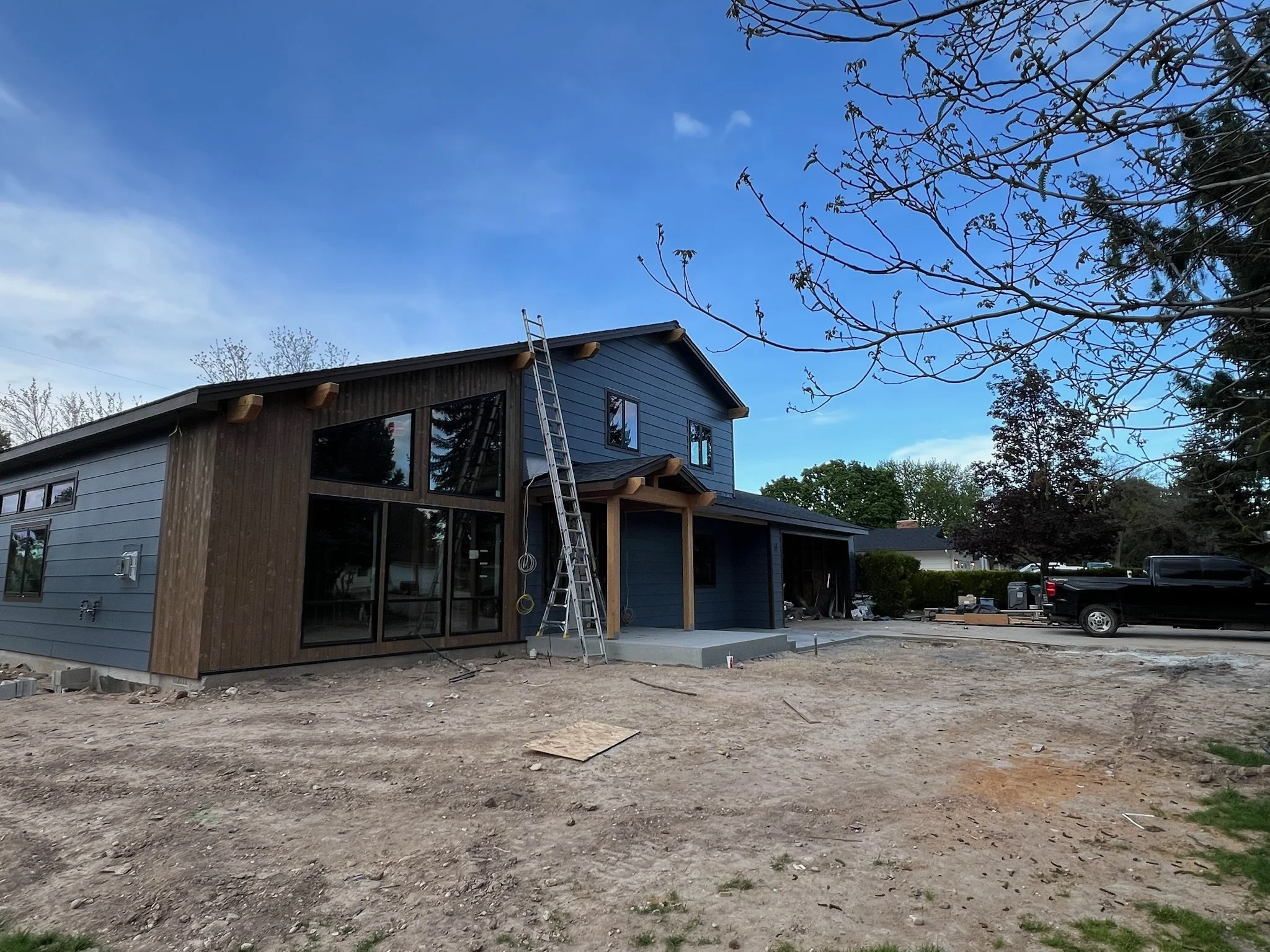 A house under construction with a ladder leaning on it, showing a mix of dark and wooden exterior siding, and a large window in the front. The yard is unfinished dirt, and there's a black truck parked to the right.