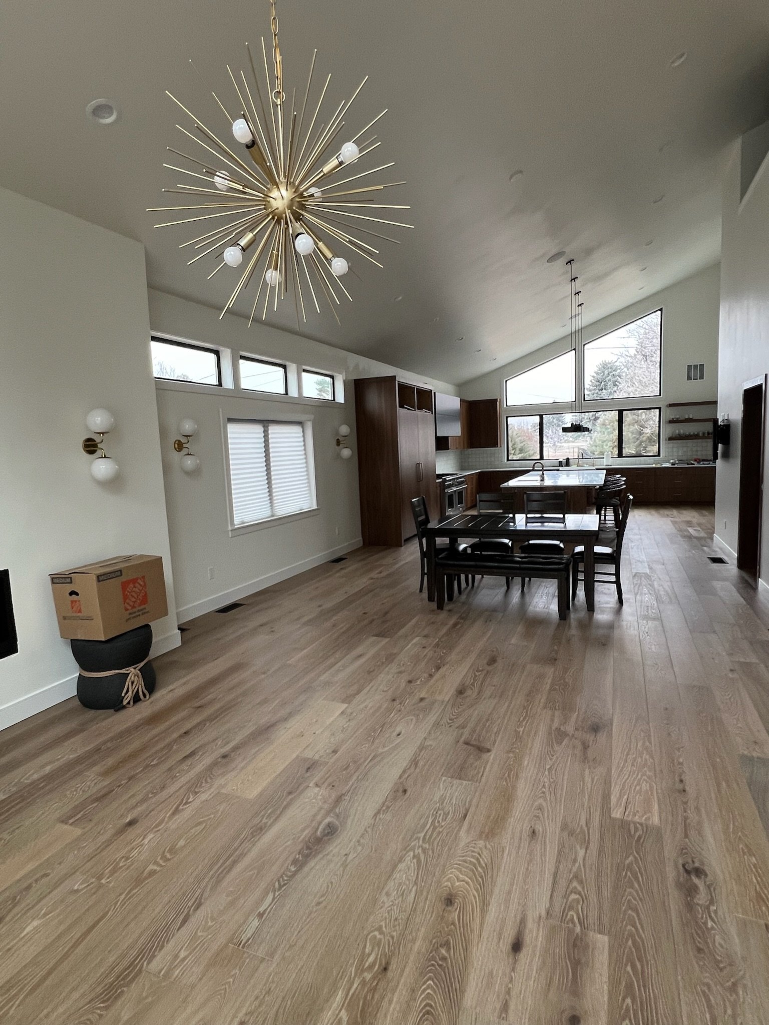 Open-concept kitchen and dining area with wooden flooring, modern chandelier, large windows, and minimal decor. There is a table with six chairs, kitchen cabinets, and a window above the sink.