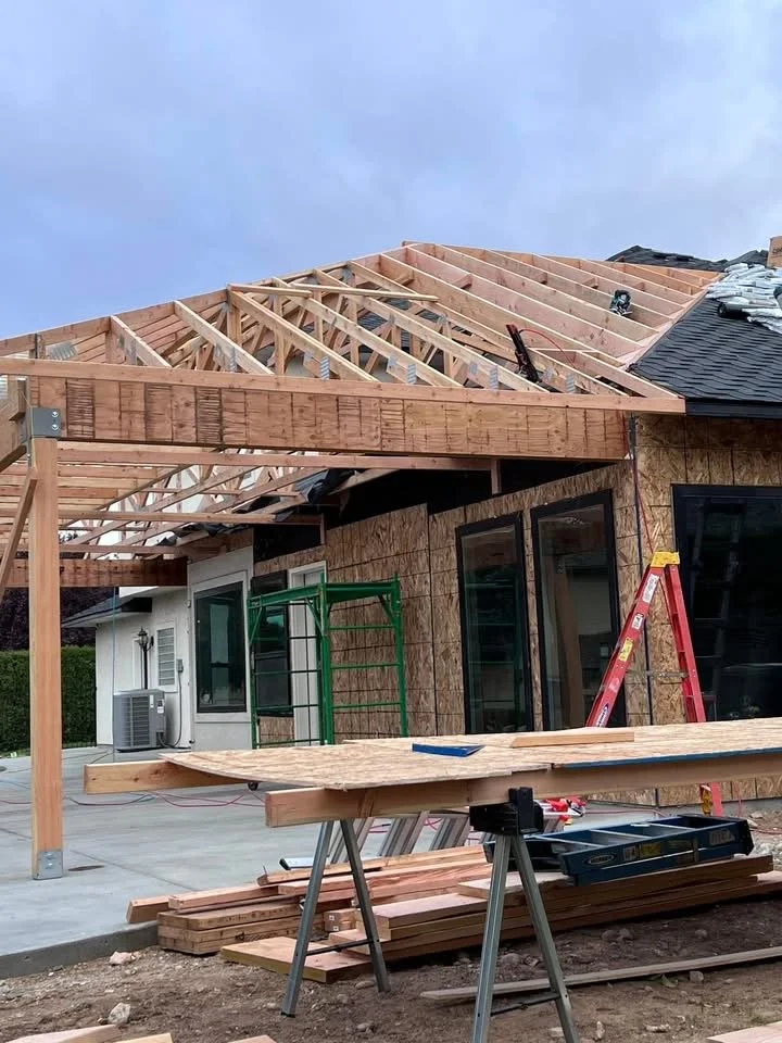 Construction site with a house undergoing roof extension or repair. Wooden framework and support beams being installed, with tools and materials on the ground.