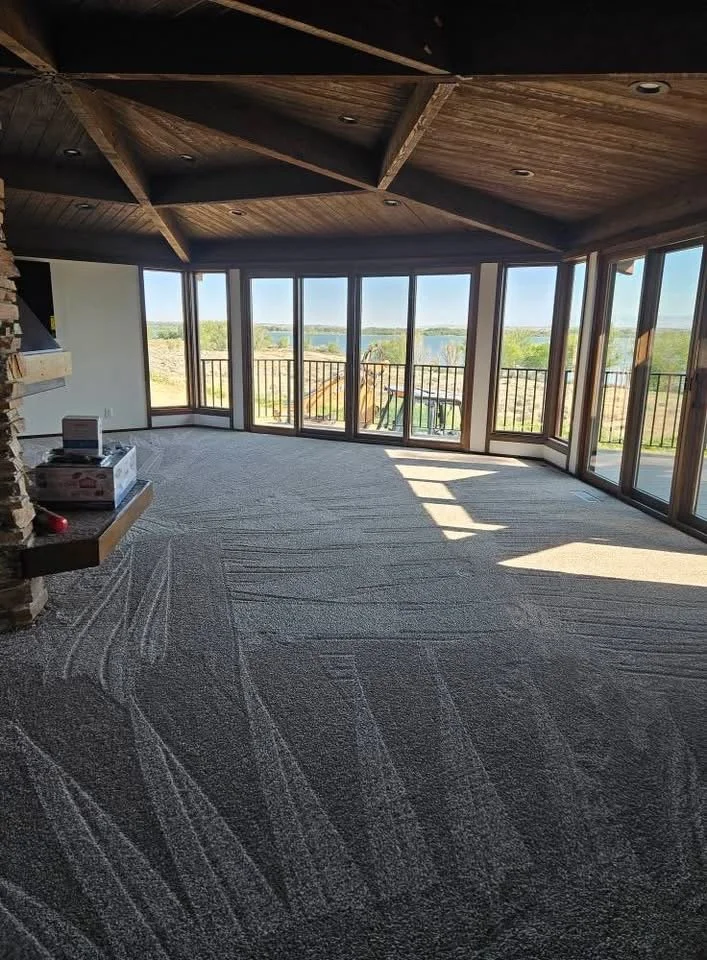 Bright, empty living room with large windows showing a scenic outdoor landscape, carpeted floor, and a wooden ceiling.